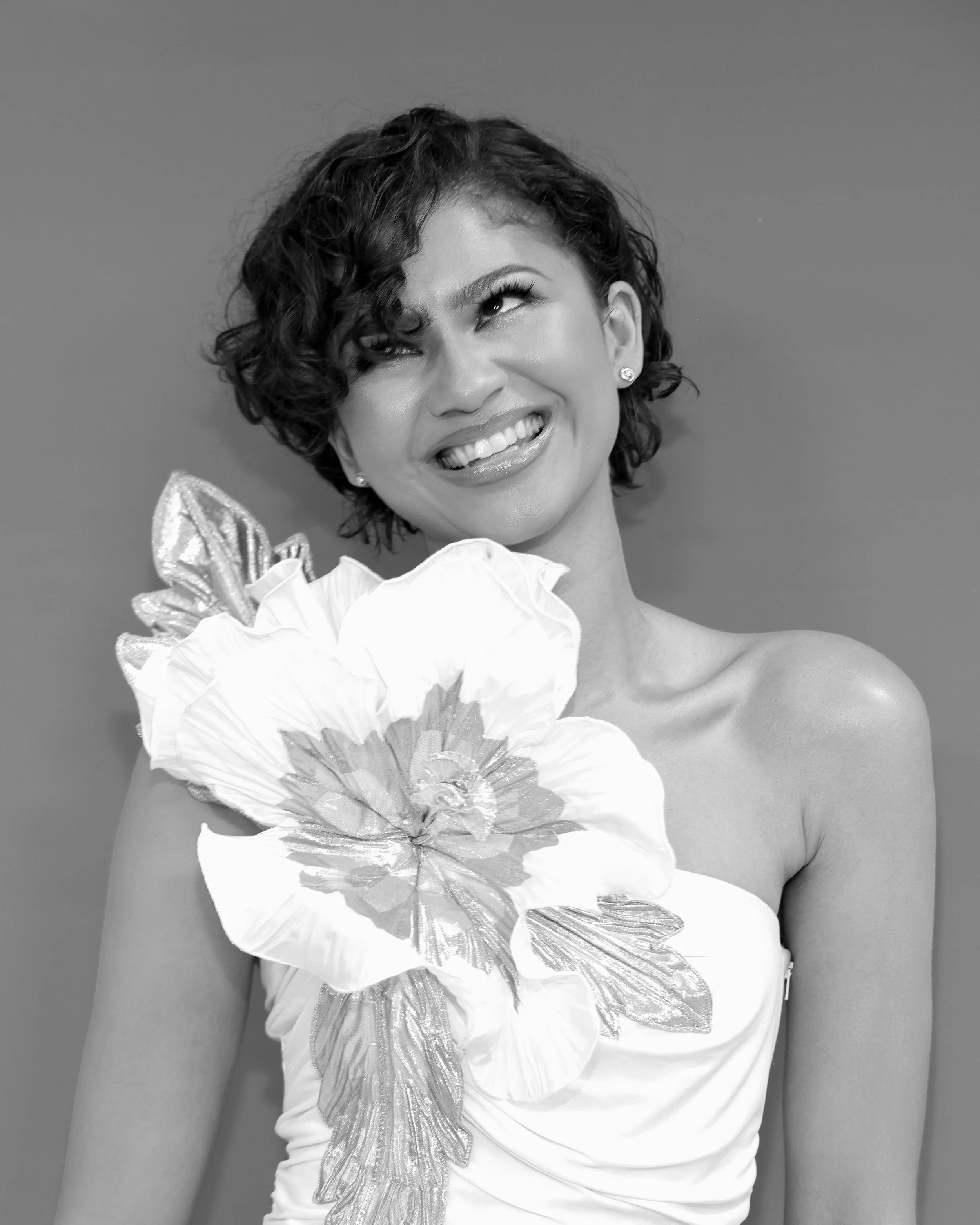 A woman with short, curly hair smiling, wearing a dress with a large floral design on the shoulder, set against a plain background.