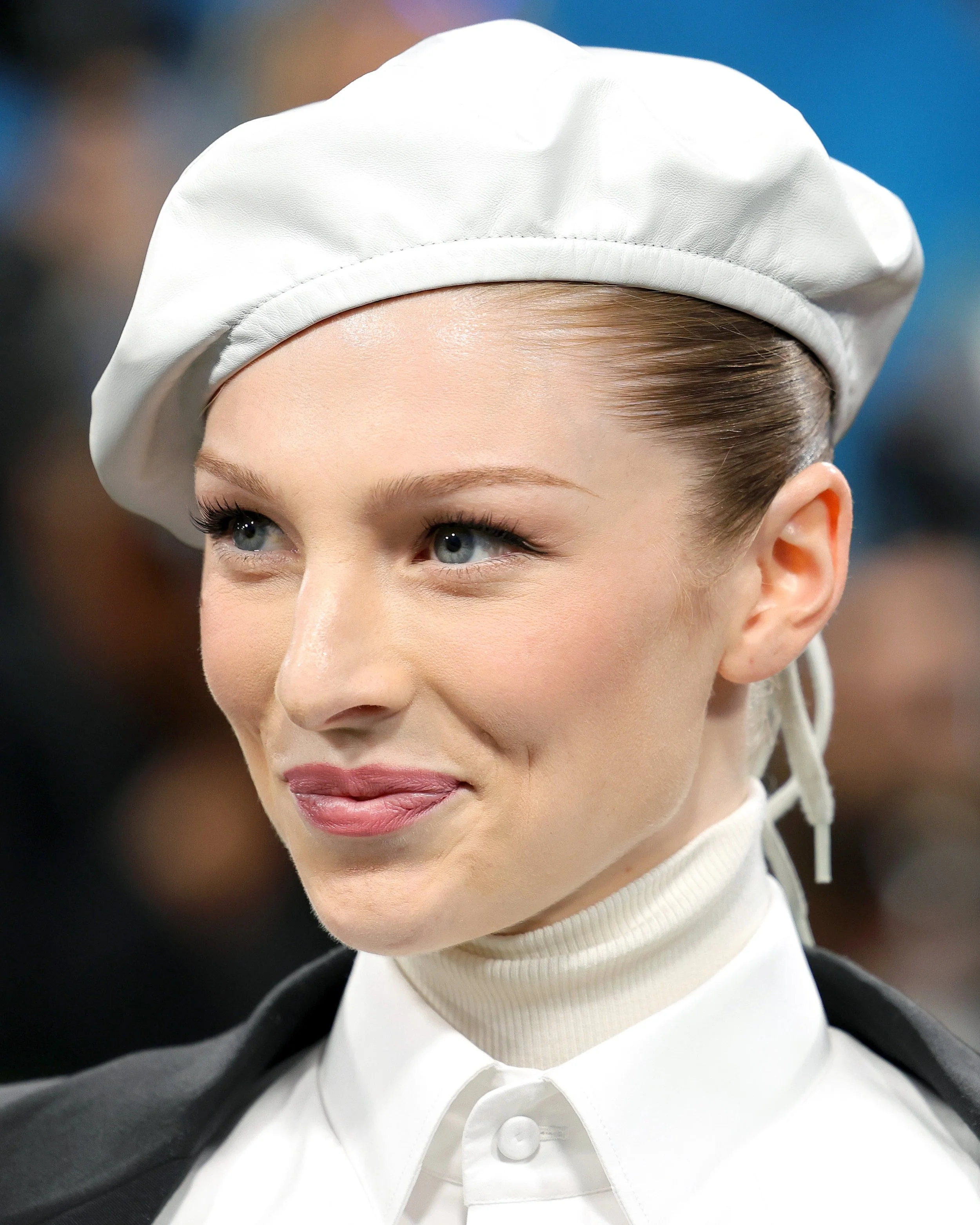 A young woman with fair skin, blue eyes, and blonde hair tied back, wearing a white chef's hat, white turtleneck, and black apron, smiling slightly.