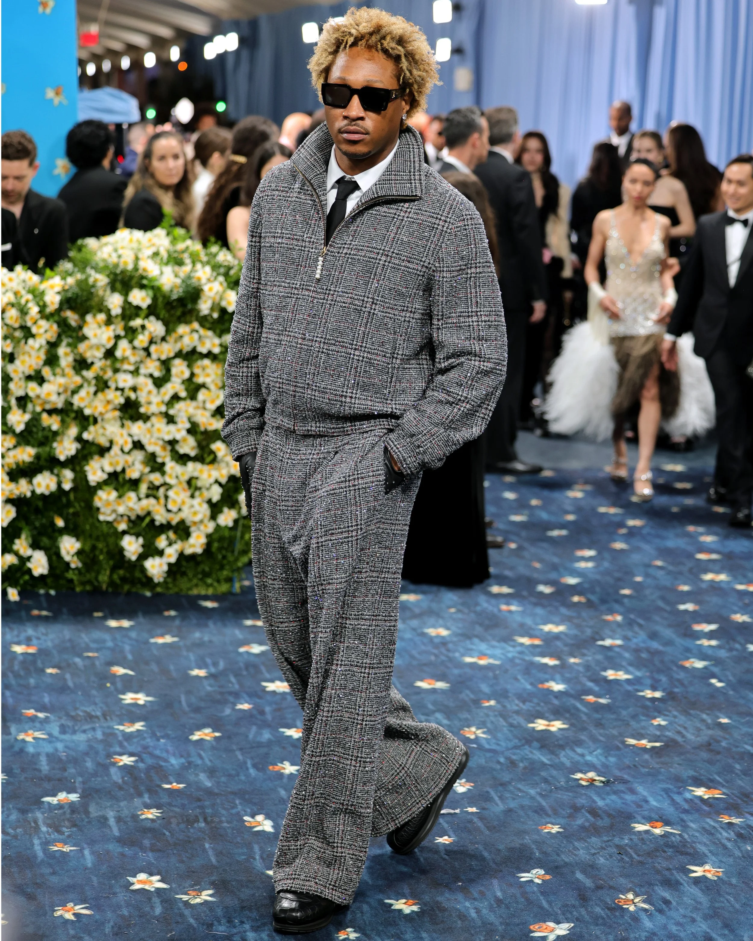 A man walking with hands in pockets, wearing a gray plaid suit, black gloves, sunglasses, and a patterned shirt and tie at a formal event, with other well-dressed people in the background.