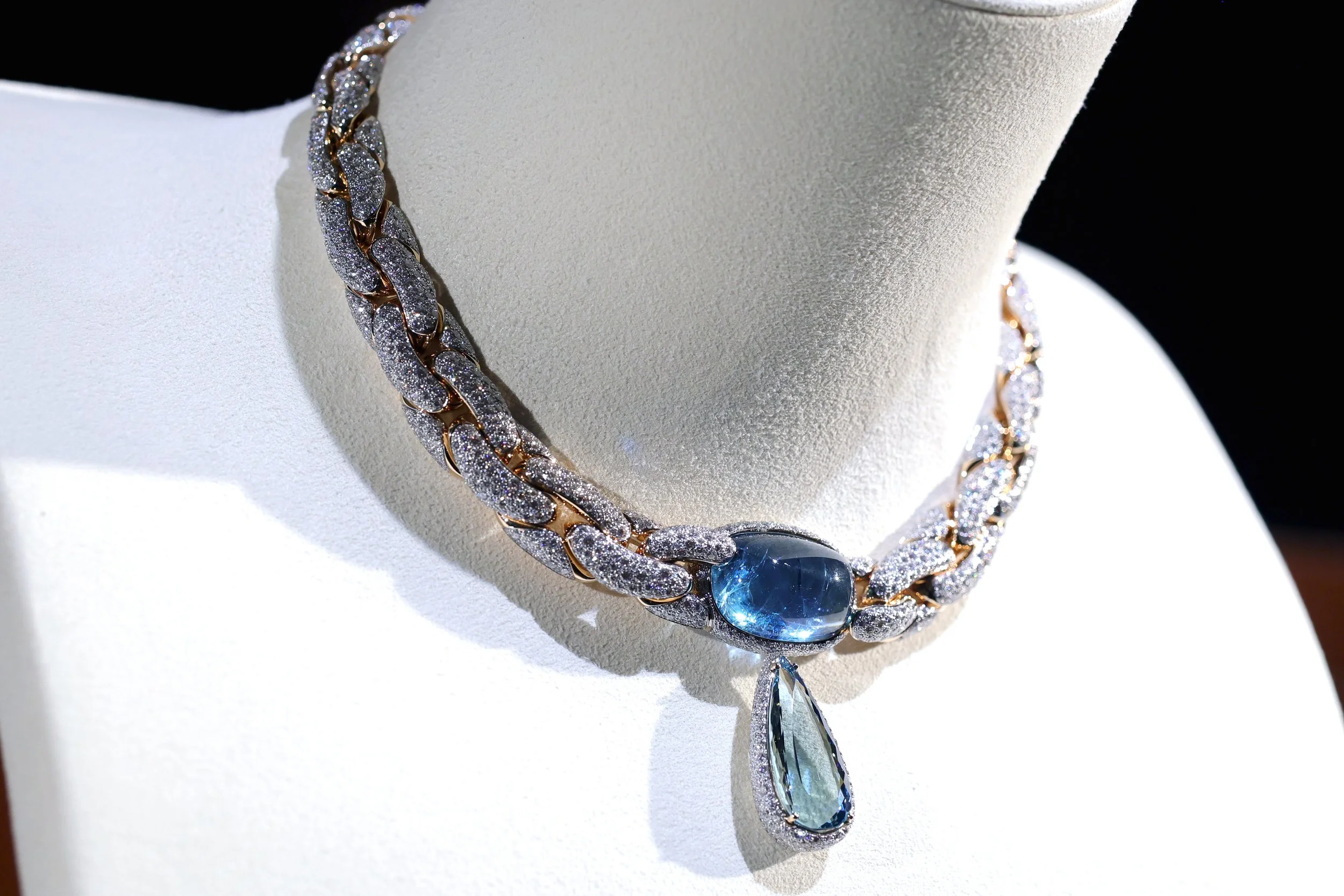 A luxurious necklace with a blue gemstone pendant and a teardrop-shaped crystal, set on a white display bust.