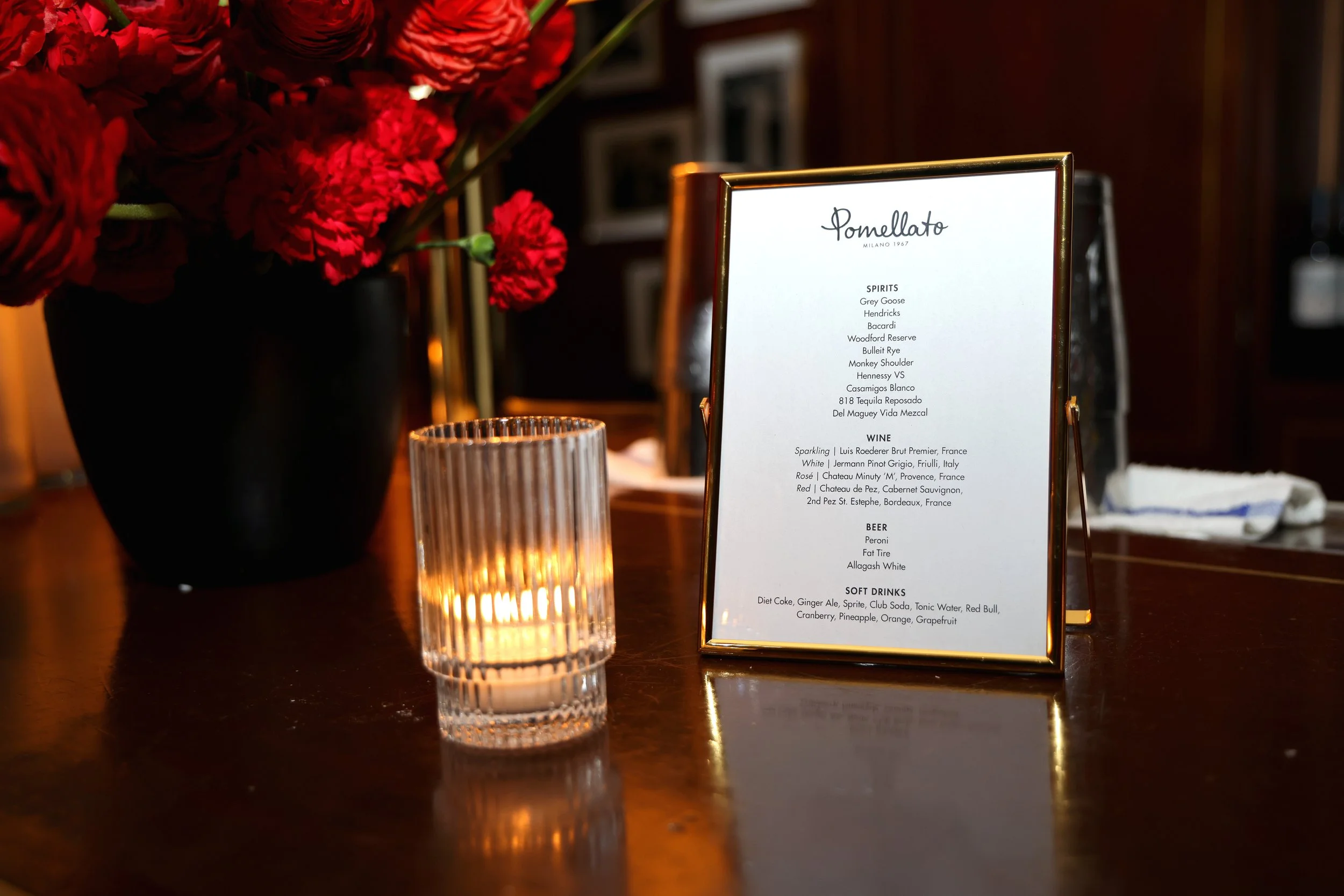 A table with a black vase filled with red flowers, a small lit candle in a ribbed glass holder, and a gold-framed menu card from Pomellato restaurant listing various spirits, wines, beers, and soft drinks.