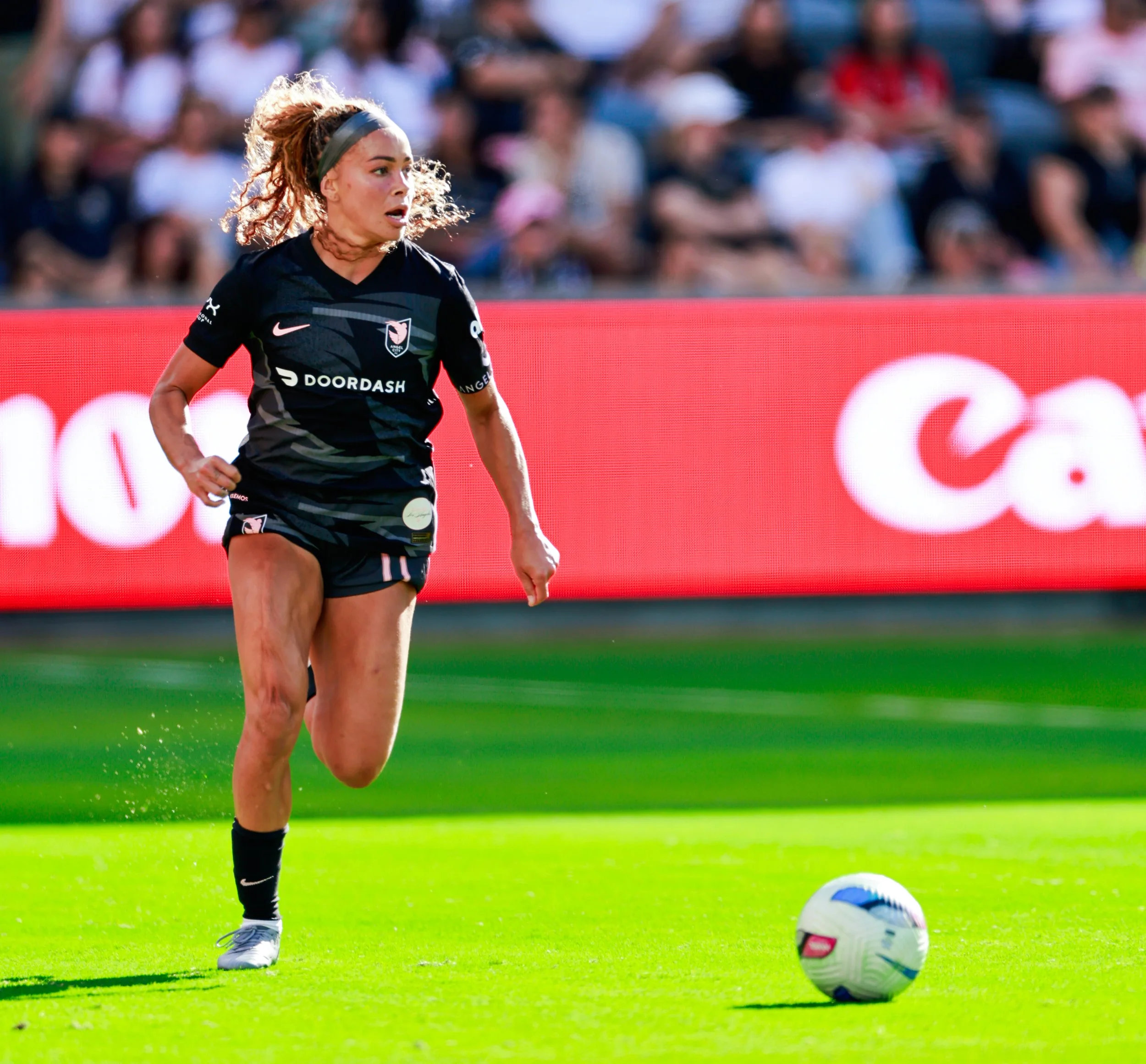 A female soccer player in a black uniform running on a soccer field before a soccer match, with a crowd in the stands behind her.