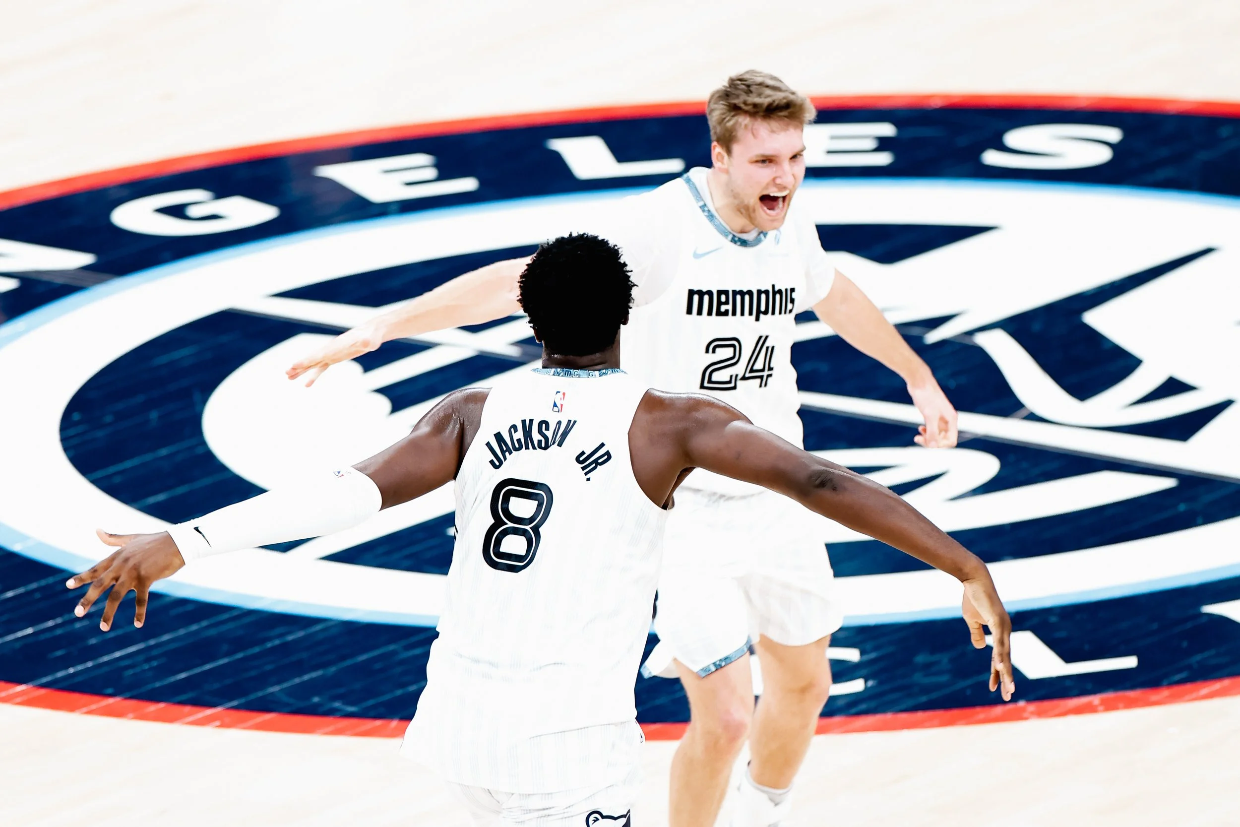 Two Memphis basketball players celebrating on the court, one with outstretched arms and the other with open arms, on a basketball court with a large Memphis logo.
