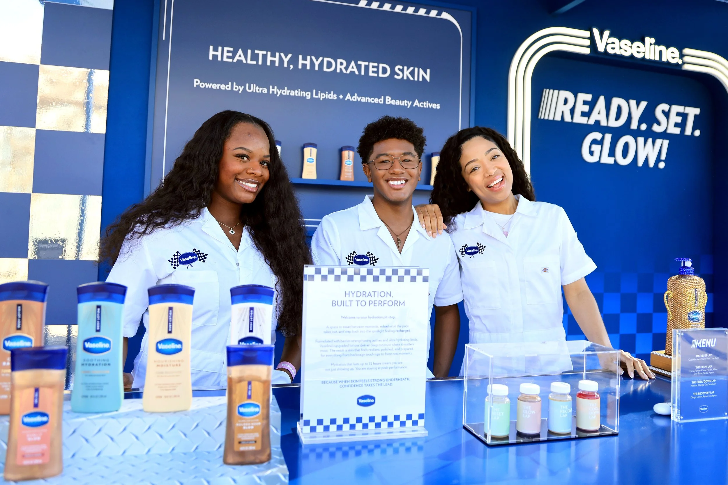 Three smiling young people in white shirts standing behind a display table at a Vaseline booth. The table has various Vaseline skincare products and informational signs, with a blue background and branding slogans like 'Healthy, Hydrated Skin' and 'R