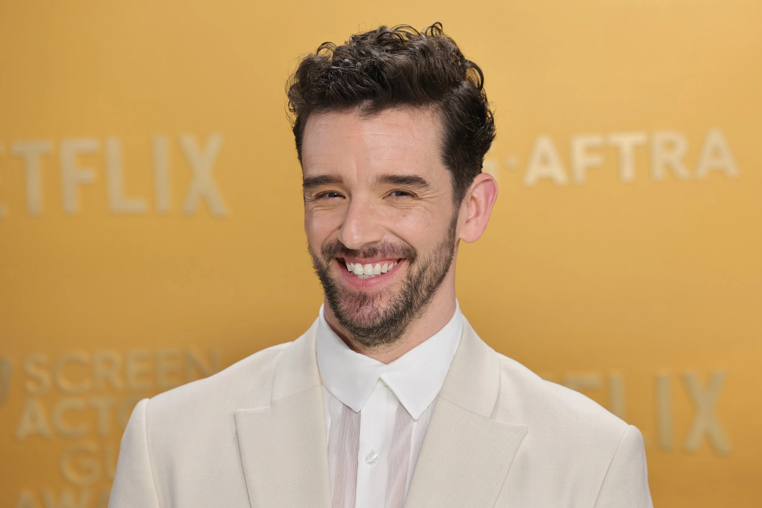 A man with dark curly hair and a beard, wearing a white suit and shirt, smiling at a yellow backdrop with the words Netflix and AFTRA.