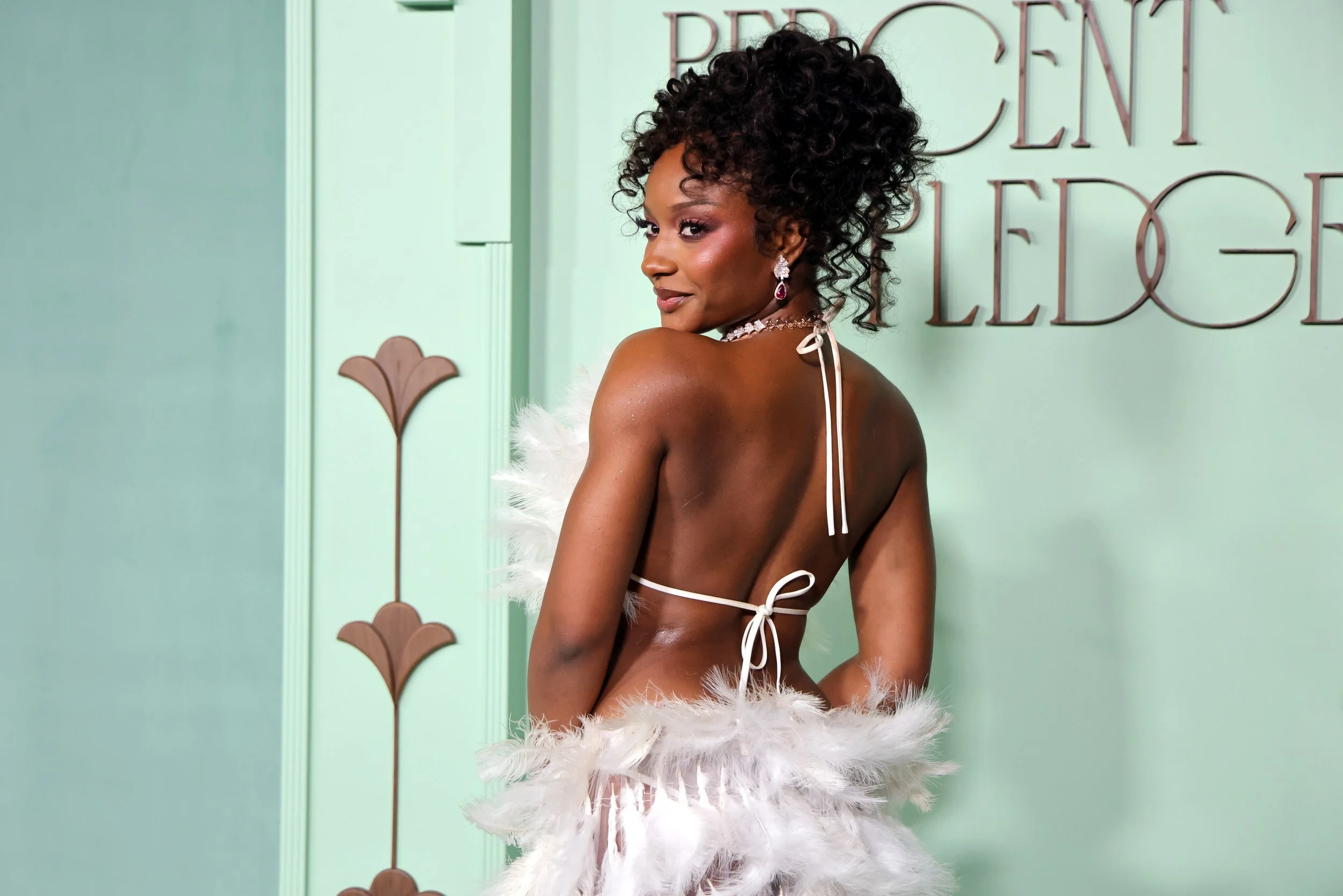 A woman with curly hair wearing a white feathered dress, looking over her shoulder with a smile, standing in front of a pastel green wall with decorative text and floral designs.