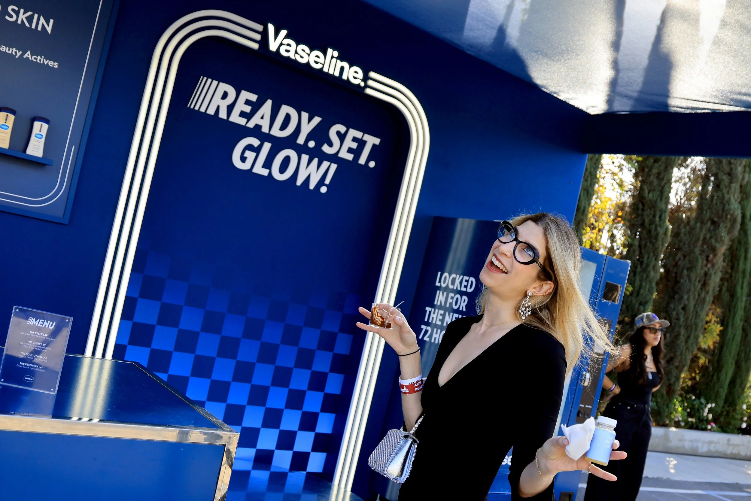 A woman with blonde hair, glasses, and earrings at a Vaseline skincare booth, holding a small jar and a snack, smiling in front of a blue display with the text 'Vaseline READY. SET. GLOW!'.