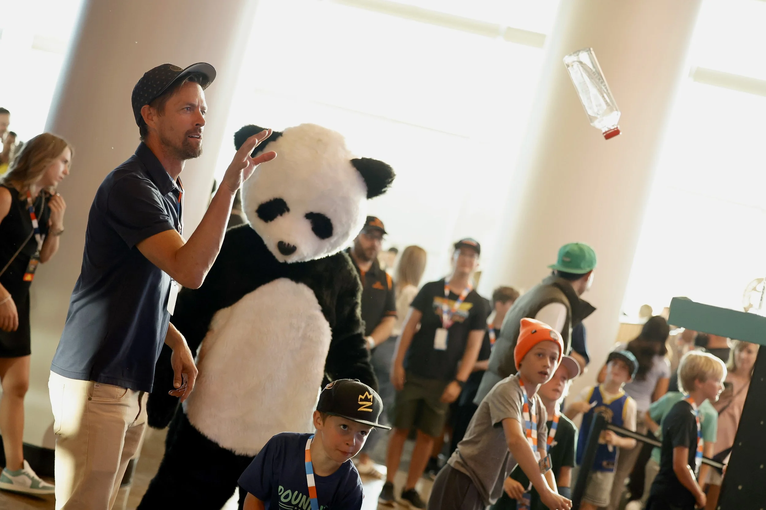 A group of children and adults in an indoor setting, with a large person in a panda costume in the center. Some children are wearing hats and medals, and a man on the left is tossing a bottle in the air.