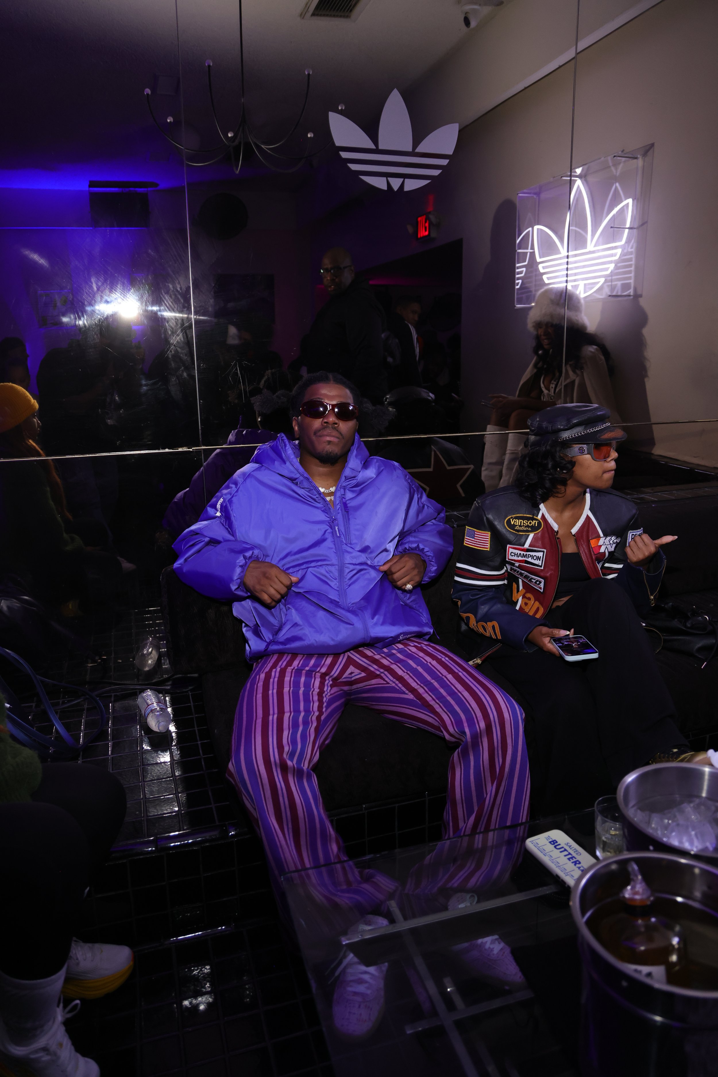 A young man in sunglasses, a purple jacket, and striped pants sitting in a lounge area at an indoor event, with a woman next to him wearing sunglasses, a black cap, and a patch-covered jacket, and a lit Adidas logo on the wall in the background.