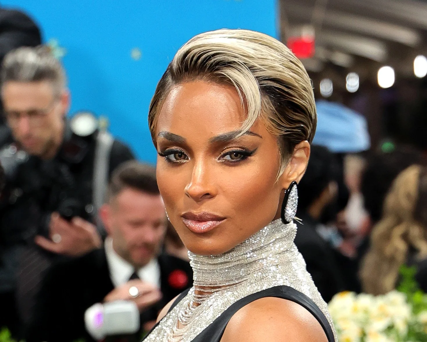 Close-up of a woman with short, styled blonde hair, wearing glamorous makeup, large earrings, and a sparkly, high-neck dress at an event with many people in the background.