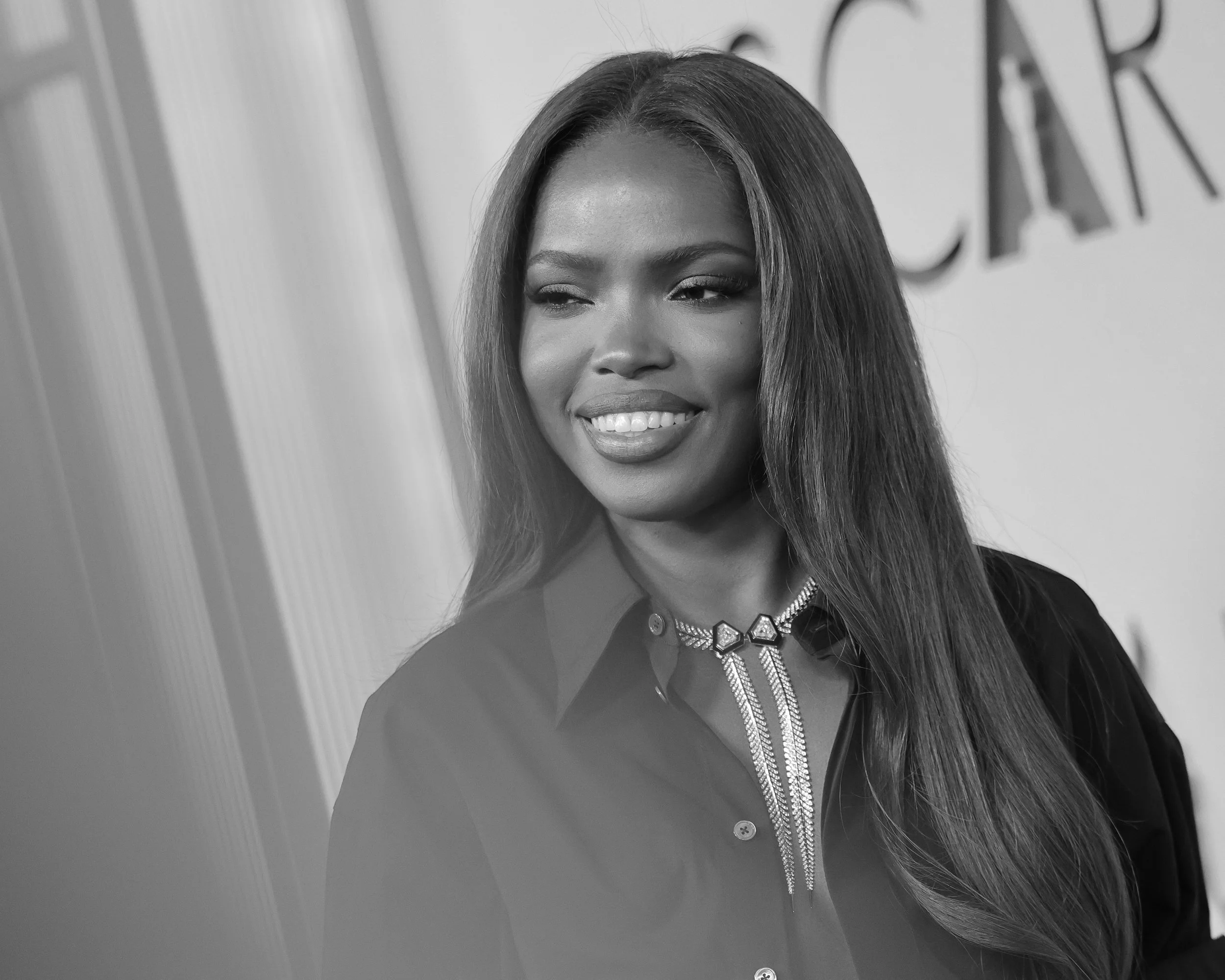 A smiling woman with long hair, wearing a dark shirt and a decorative necklace, standing indoors in front of a wall with partial text visible.