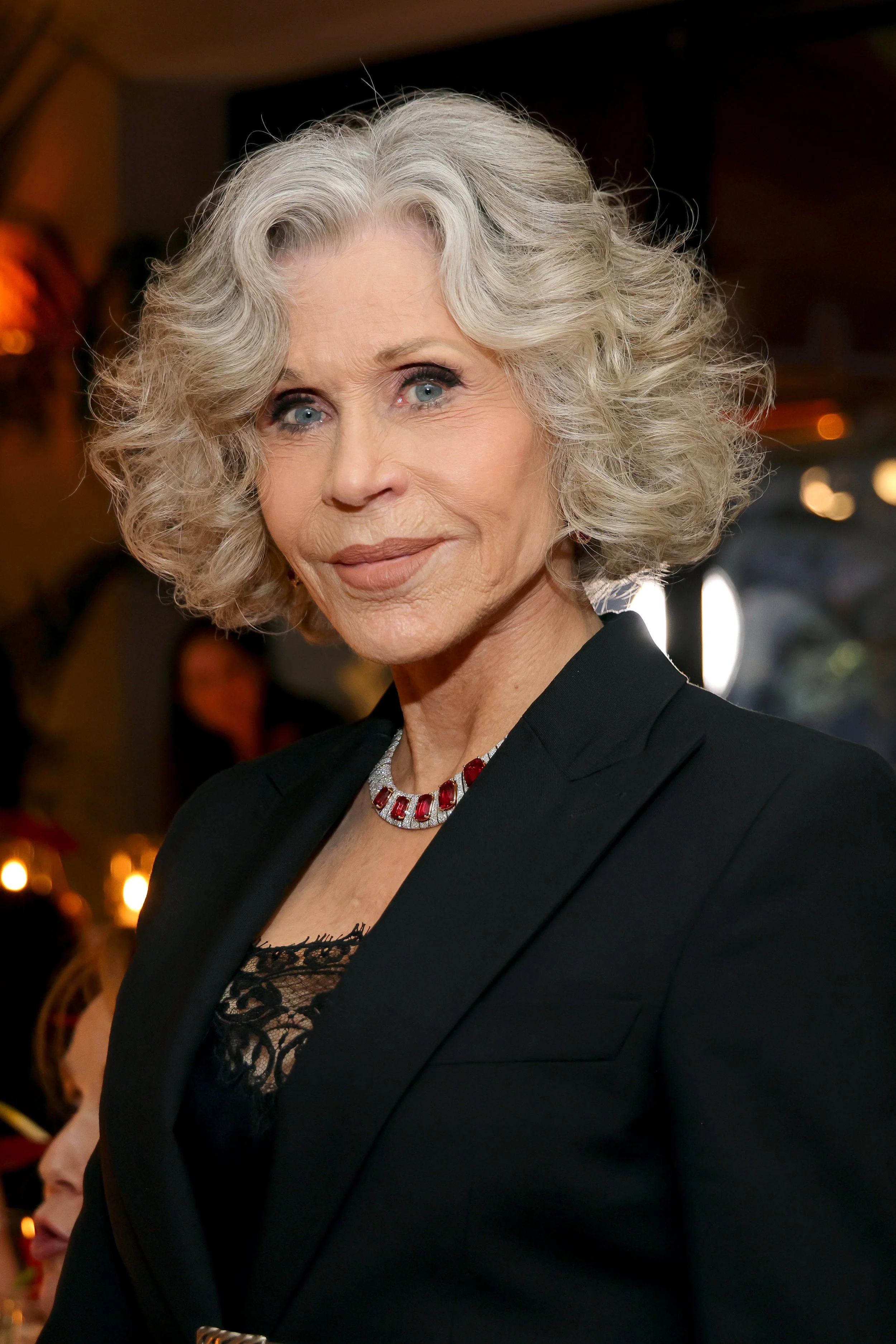 An older woman with curly gray hair, wearing a black blazer, lace top, and red jewelry, posing at an event with warm lighting and blurred background.