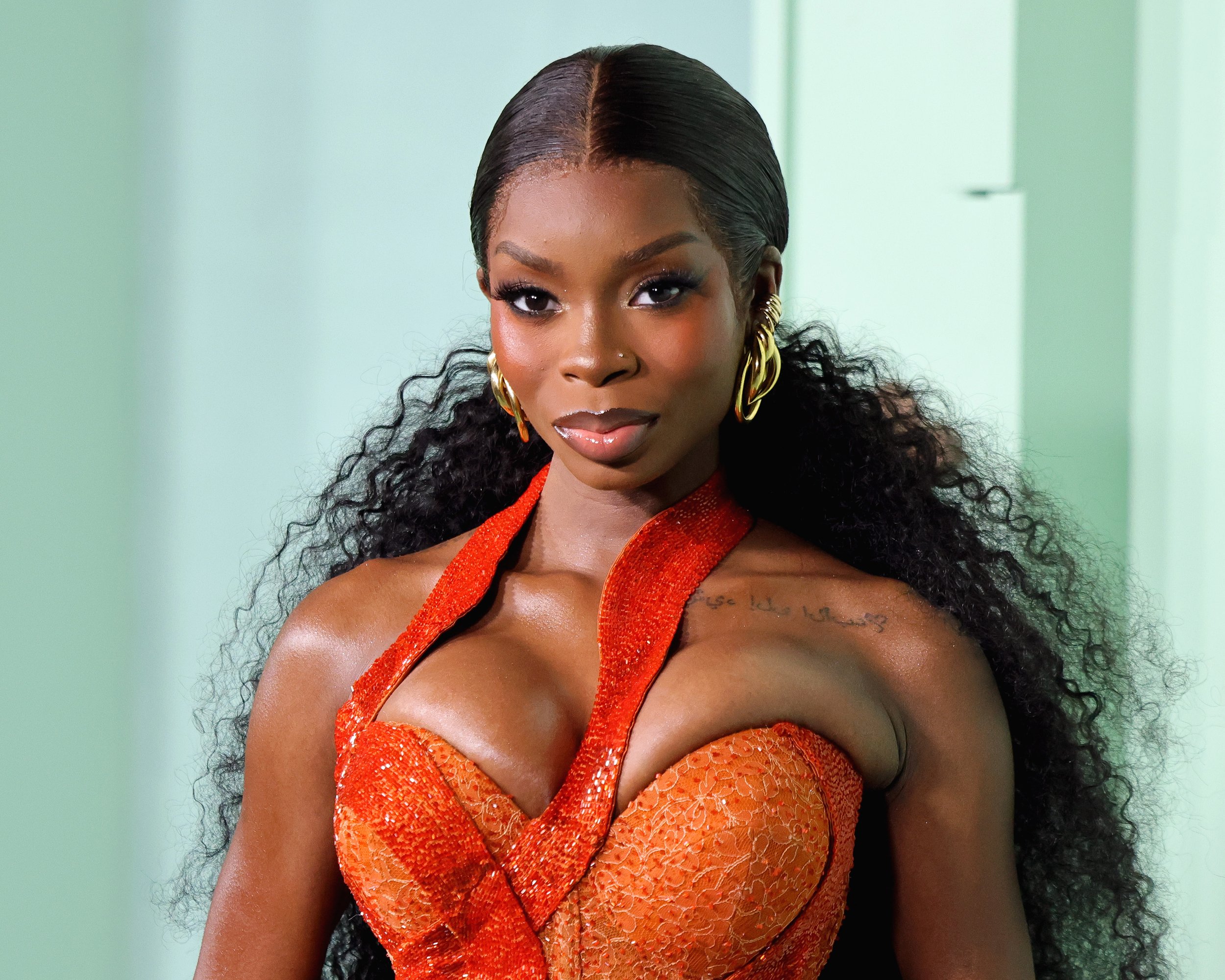 A woman with dark curly hair wearing large gold earrings and a red-orange lace dress with a deep neckline, looking directly at the camera.