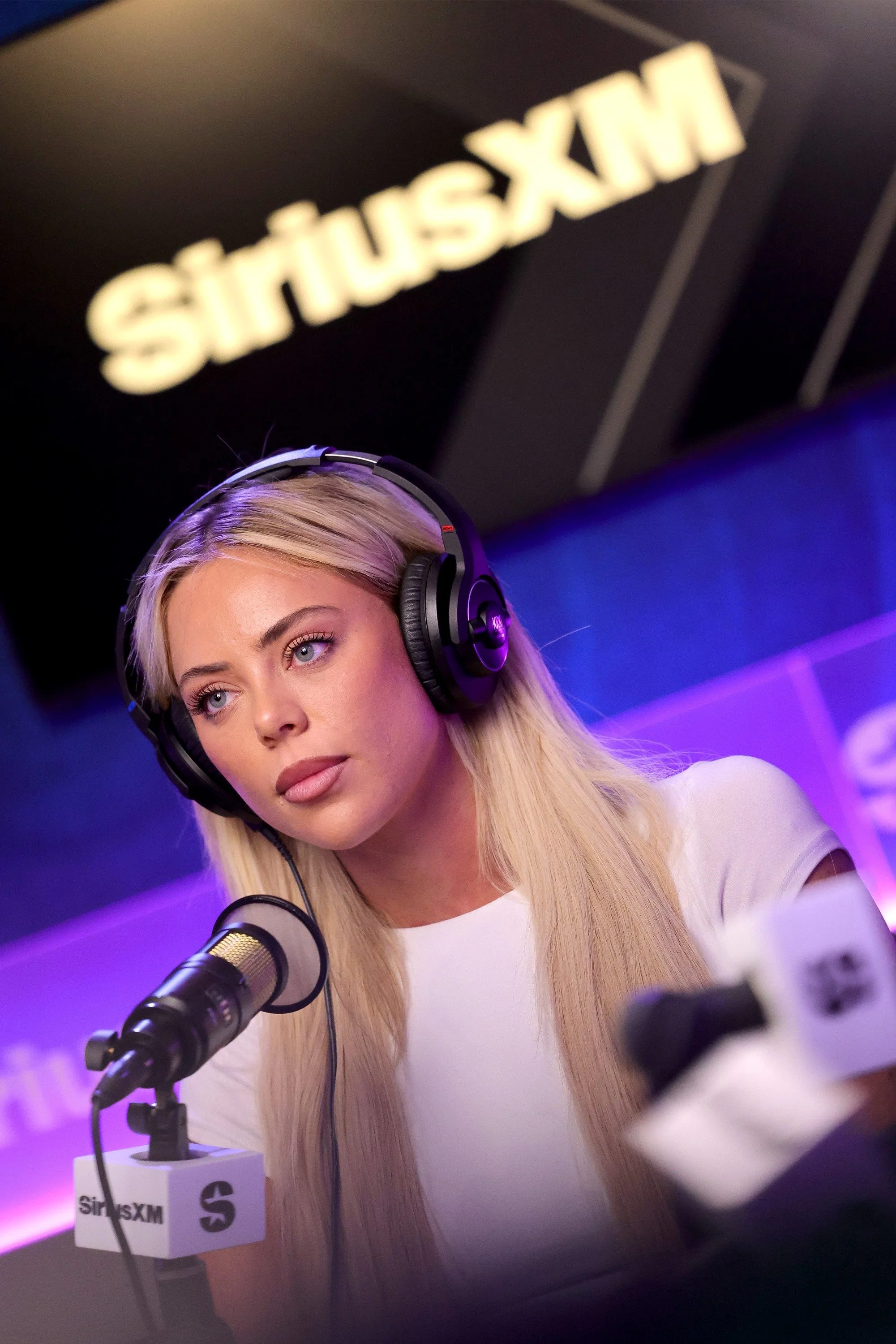 A woman with long blonde hair wearing headphones and speaking into a microphone at a SiriusXM radio station.