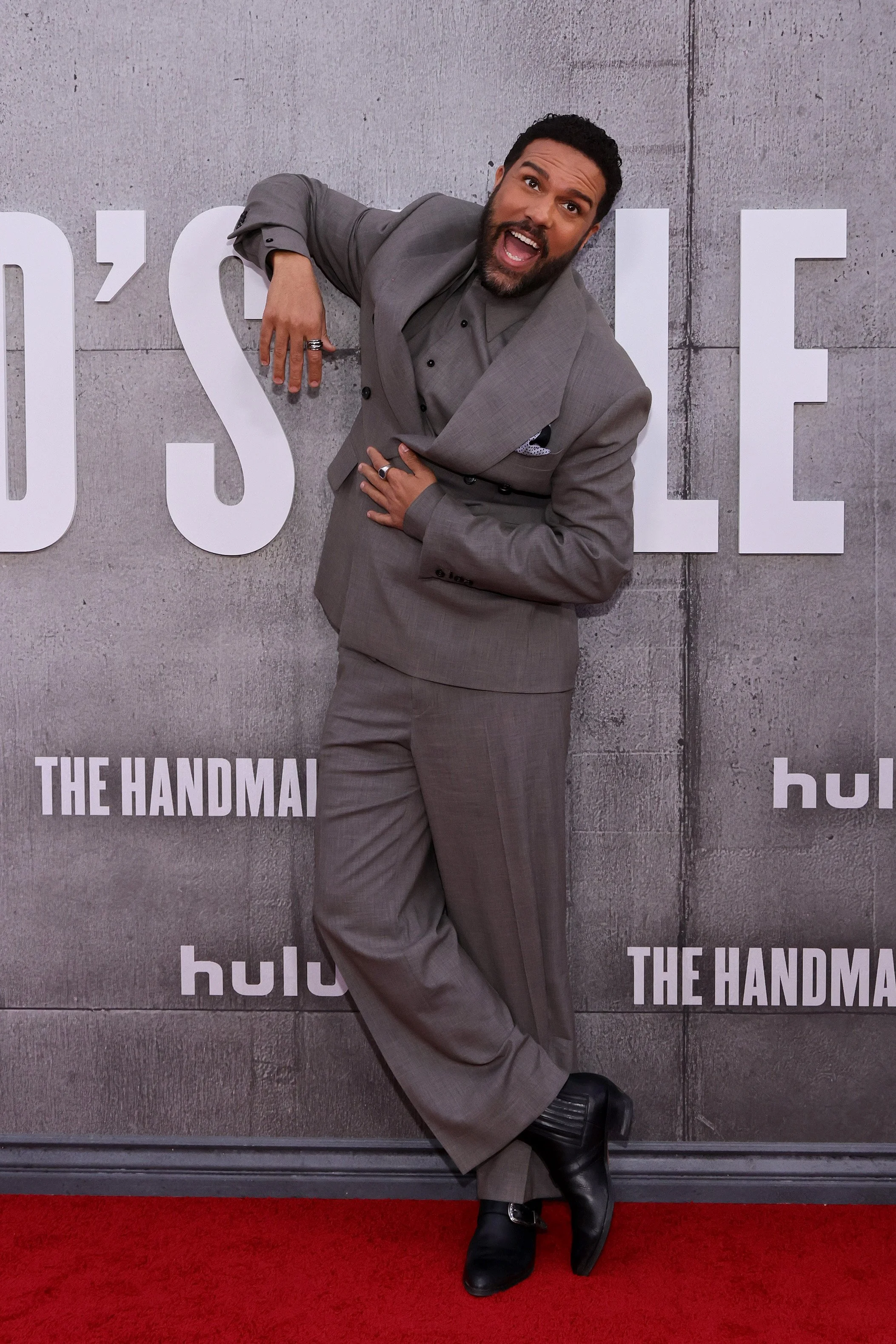A man in a gray suit posing on a red carpet with a playful expression, against a backdrop promoting the show 'The Handmaid's Tale' on Hulu.