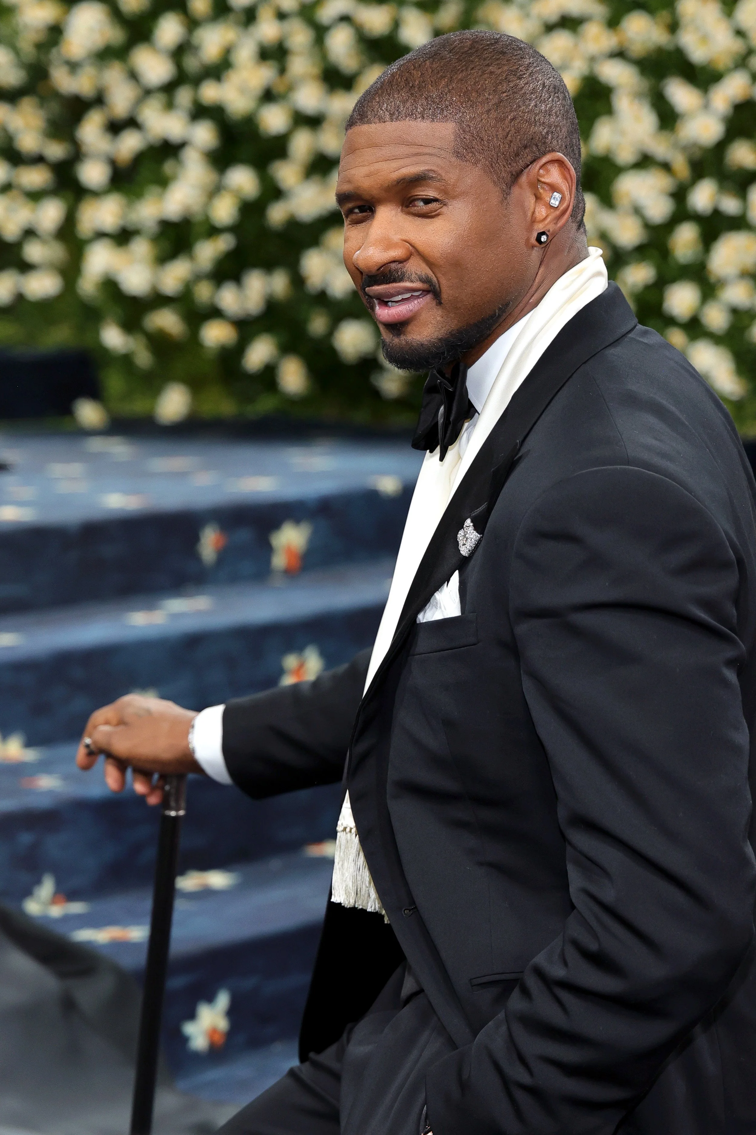 A man in a black tuxedo and bow tie standing near a staircase with floral decoration, holding a cane, outdoors.