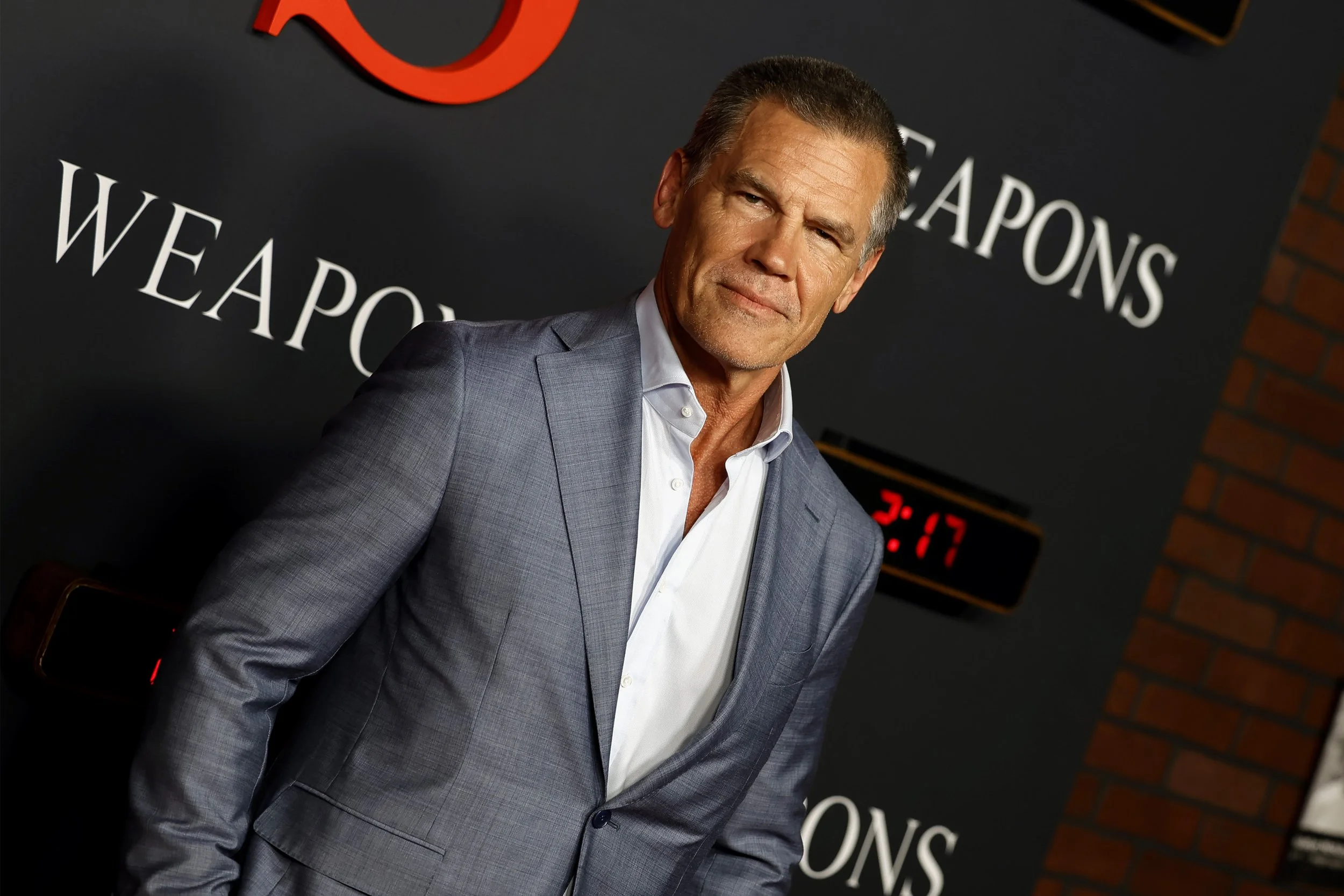 A man in a gray suit and white shirt standing in front of a black background with the word "WEAPONS" and a red digital clock showing "2:17."
