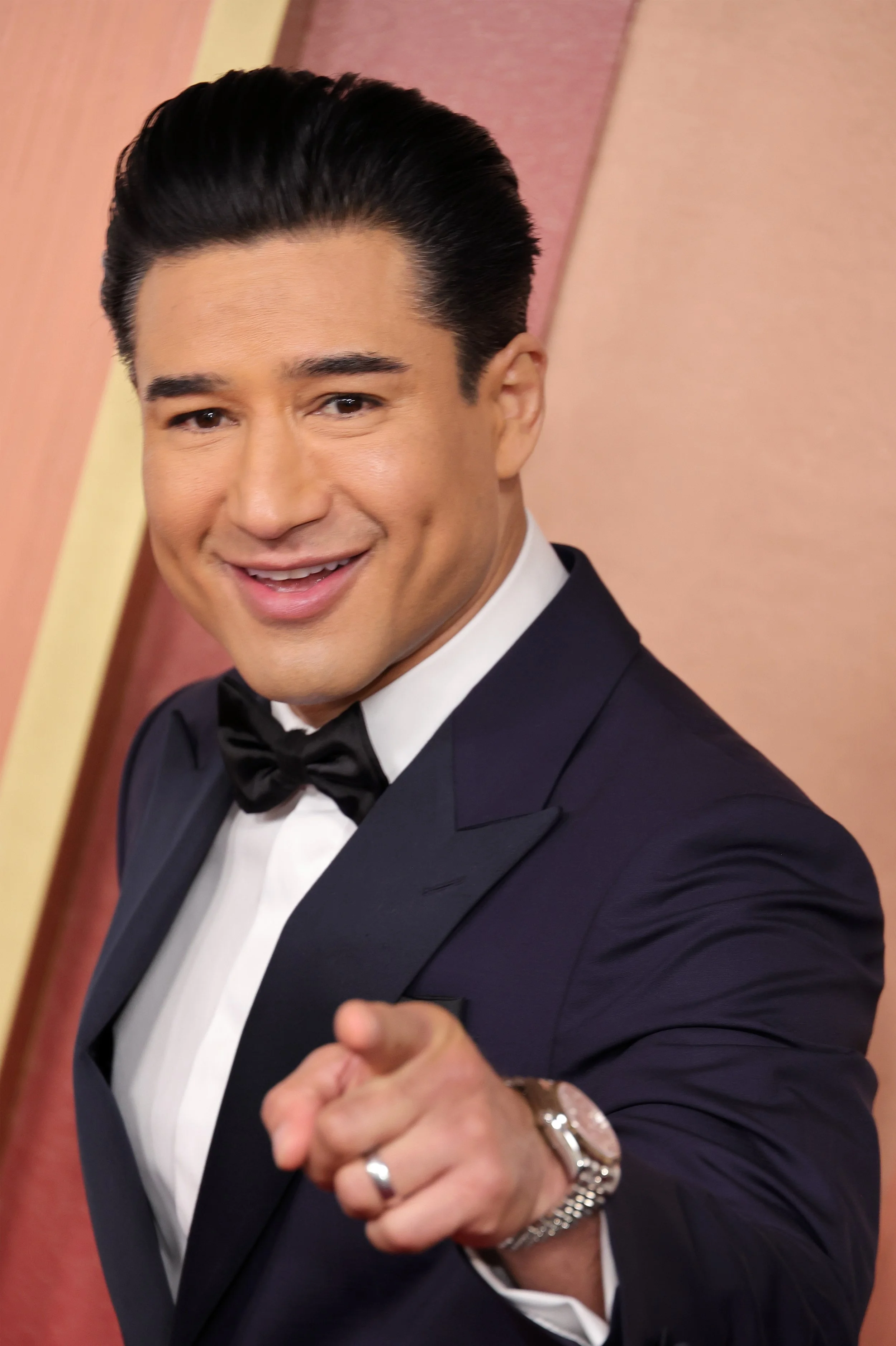 A smiling man in a tuxedo and bow tie pointing at the camera, standing against a colorful background.