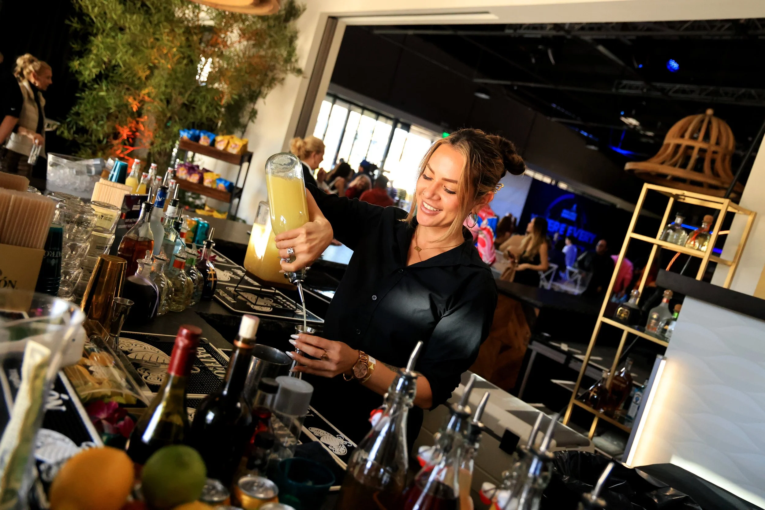 A smiling woman in black shirt makes a cocktail at a bar with various bottles and fruits, in a busy indoor event space.