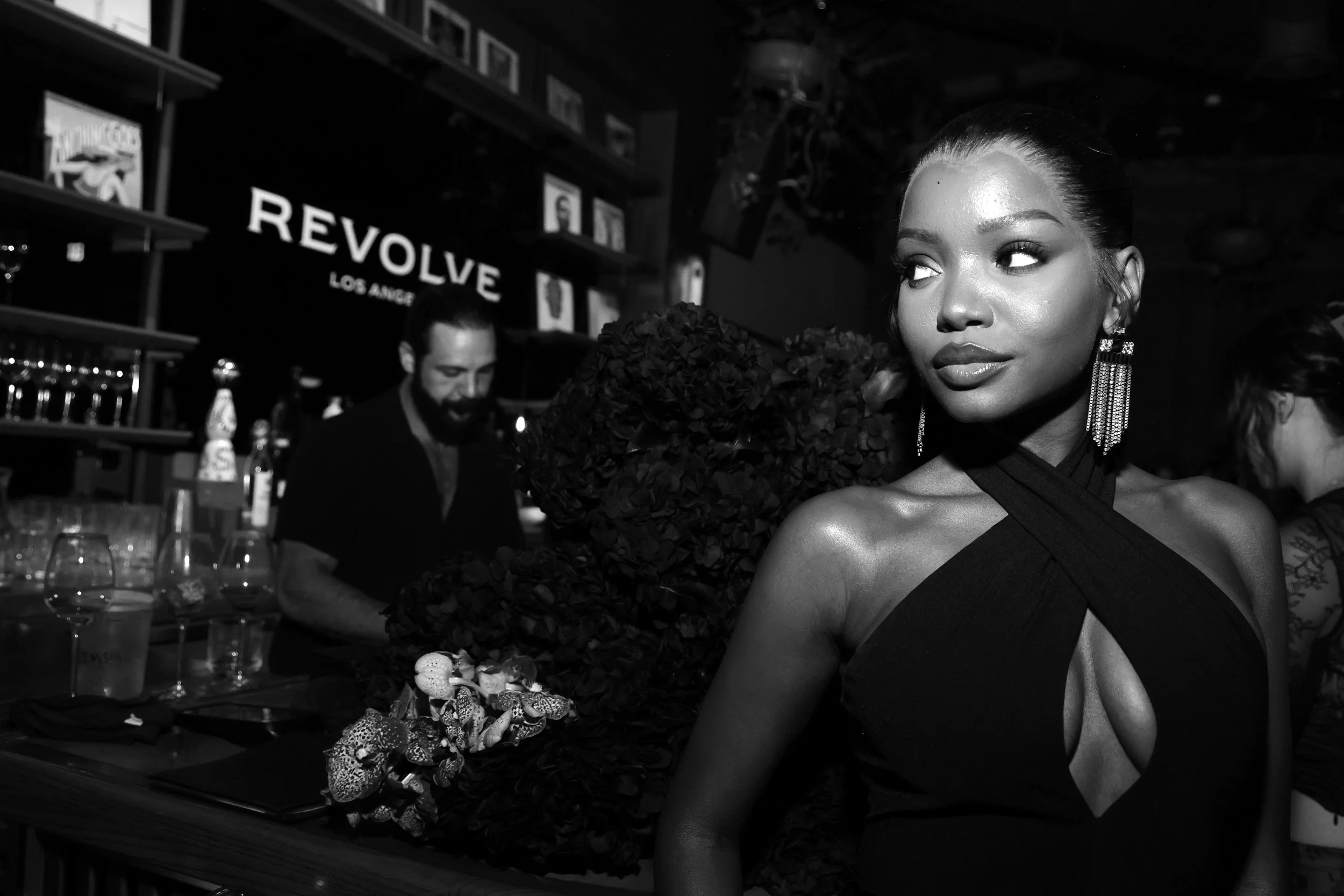 A woman with styled hair and large earrings at a bar or event, with a man behind her preparing drinks and a sign that reads "REVOLVE Los Angeles" in the background.
