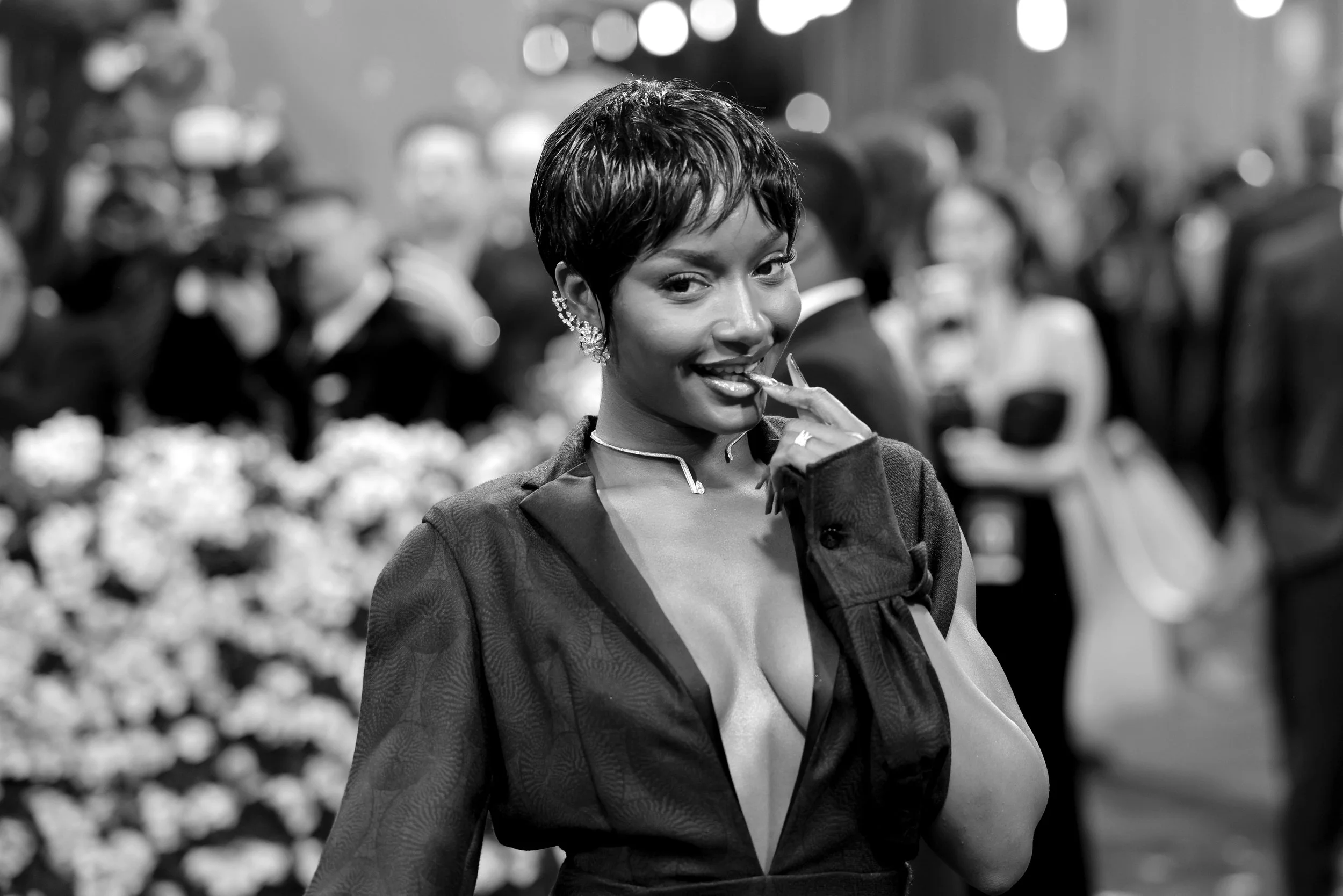 A woman with short hair, wearing earrings, a deep V-neck dress, and a jacket, looking at the camera and smiling while touching her lips, at a social gathering with blurred people and flowers in the background.