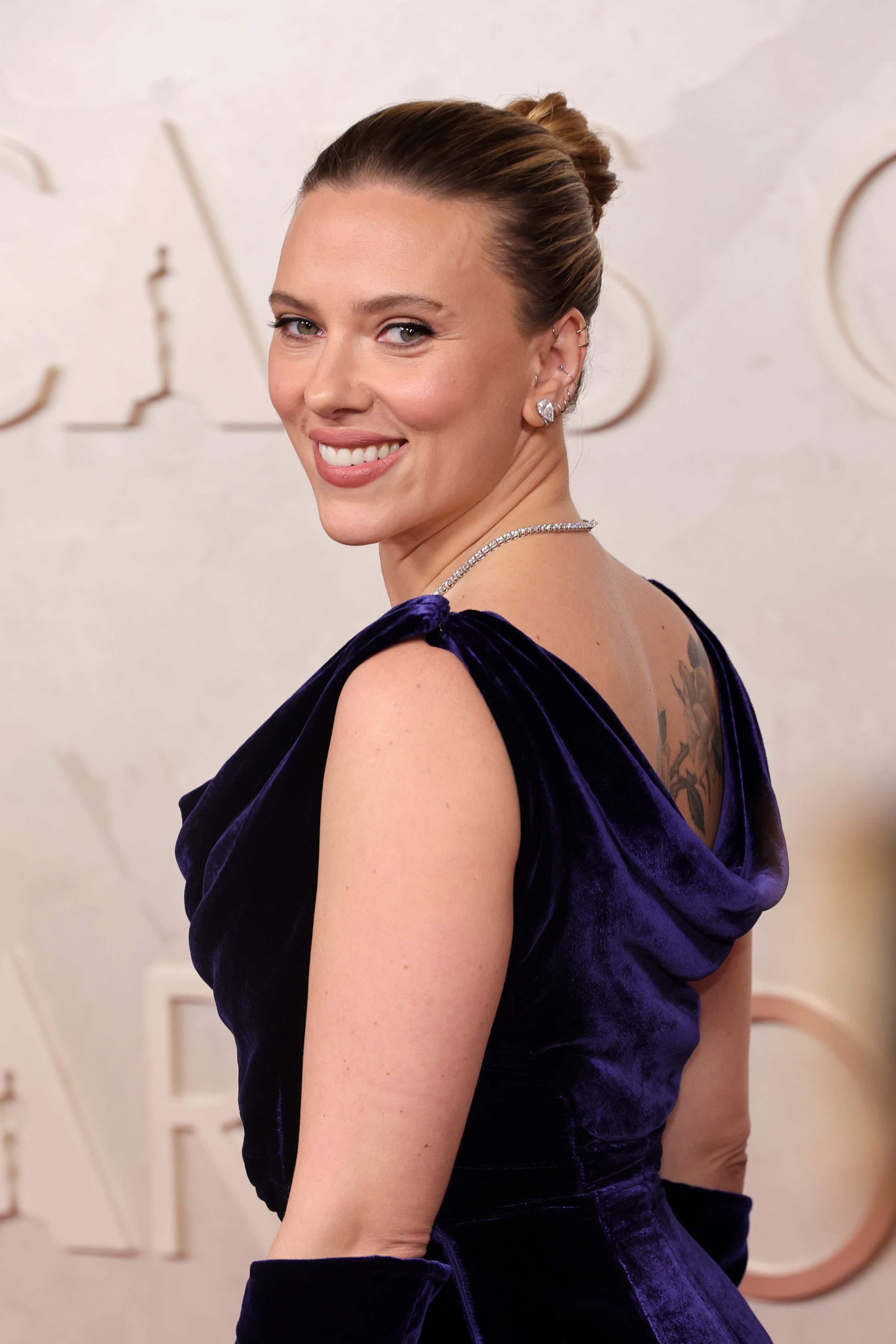 A woman in a dark purple velvet dress with a tattoo visible on her upper back, smiling at the camera, with an elegant updo hairstyle and jewelry.