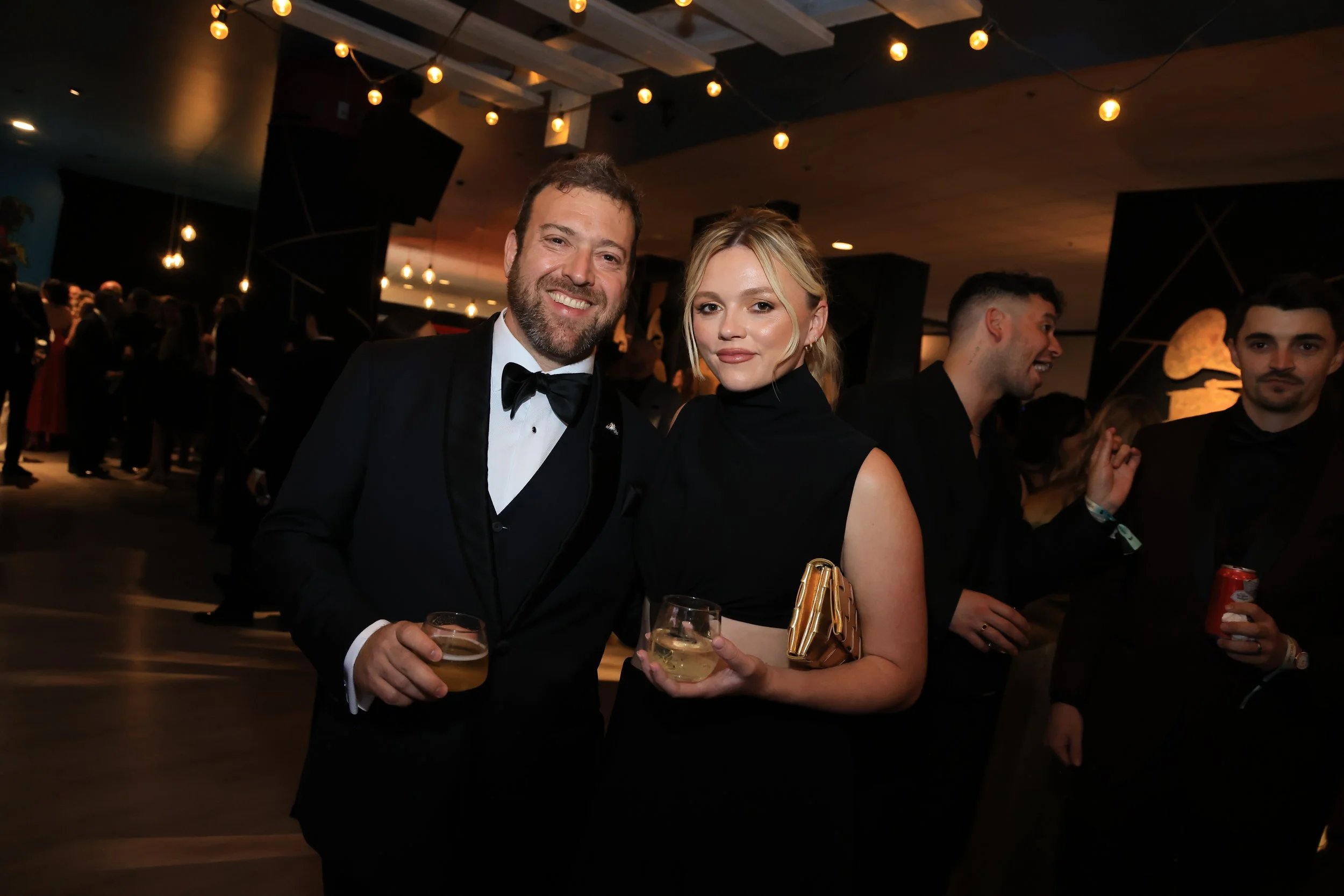 A man in a tuxedo and a woman in a black dress posing together at a formal event, holding drinks in a crowded, dimly lit room with string lights overhead.