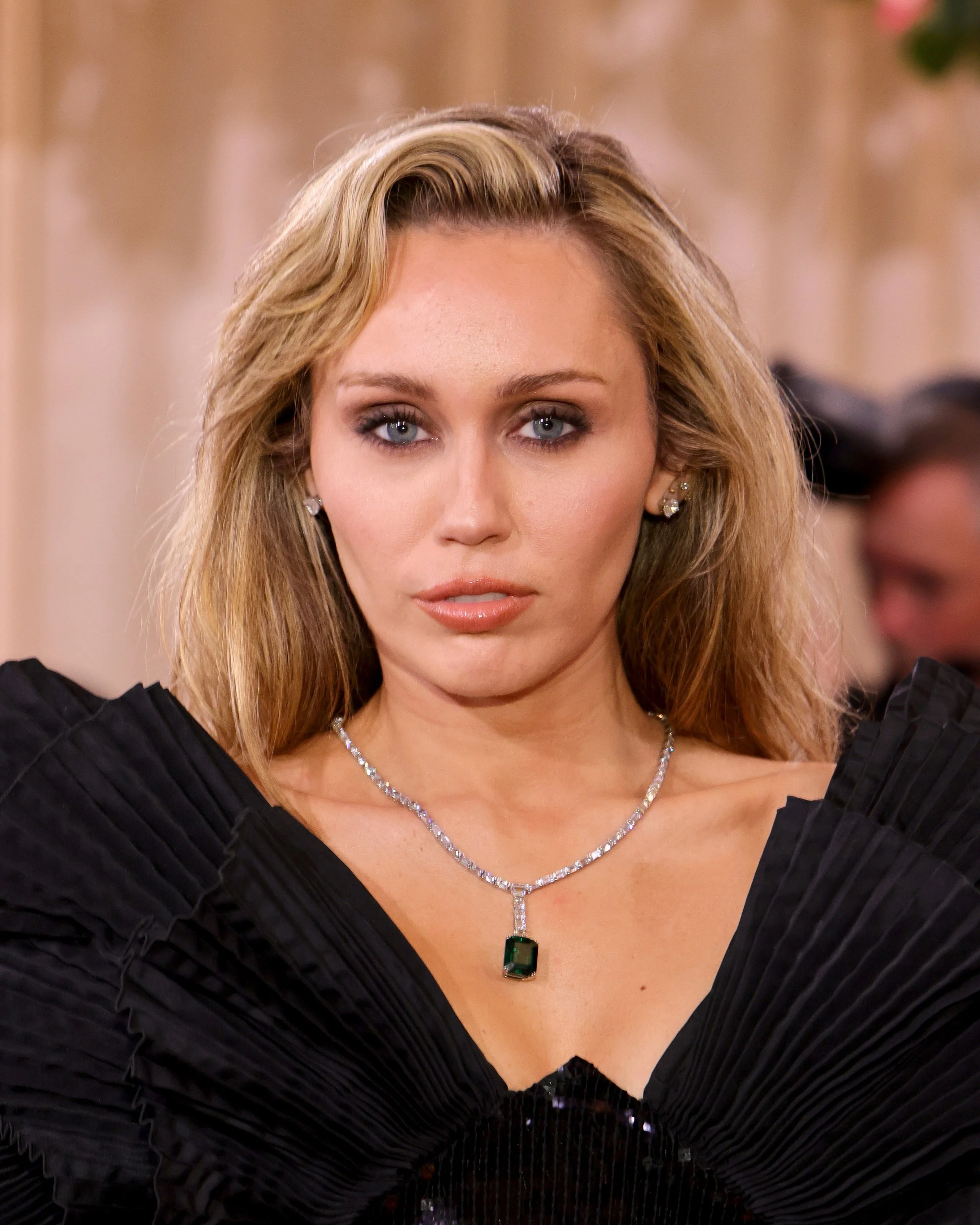 A woman with blonde hair and blue eyes wearing a black dress and a sparkling necklace with a large emerald pendant.