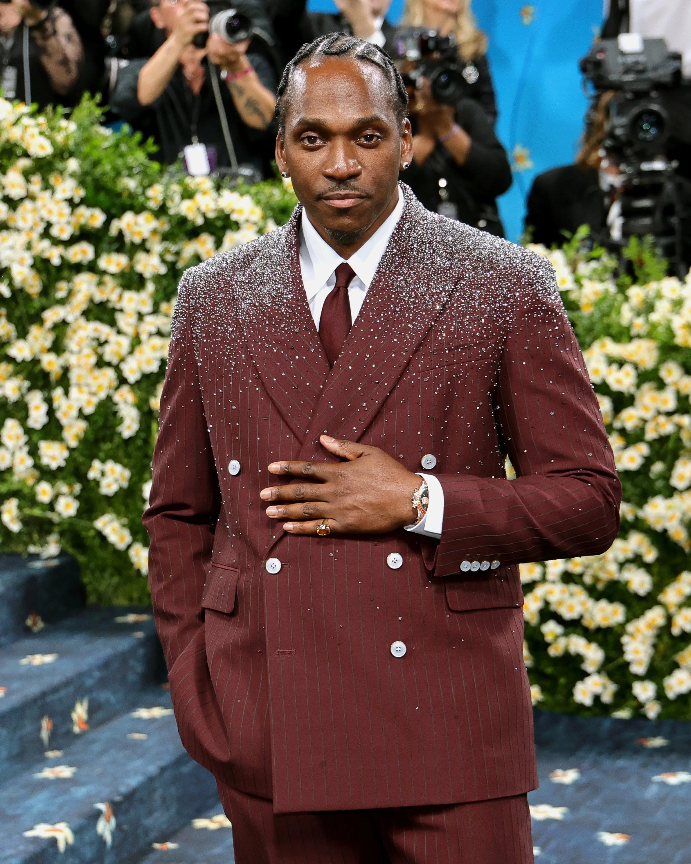 A man in a burgundy double-breasted suit with rhinestone embellishments, white shirt, and burgundy tie, standing with one hand over his chest during a formal event, surrounded by photographers and floral decorations in the background.