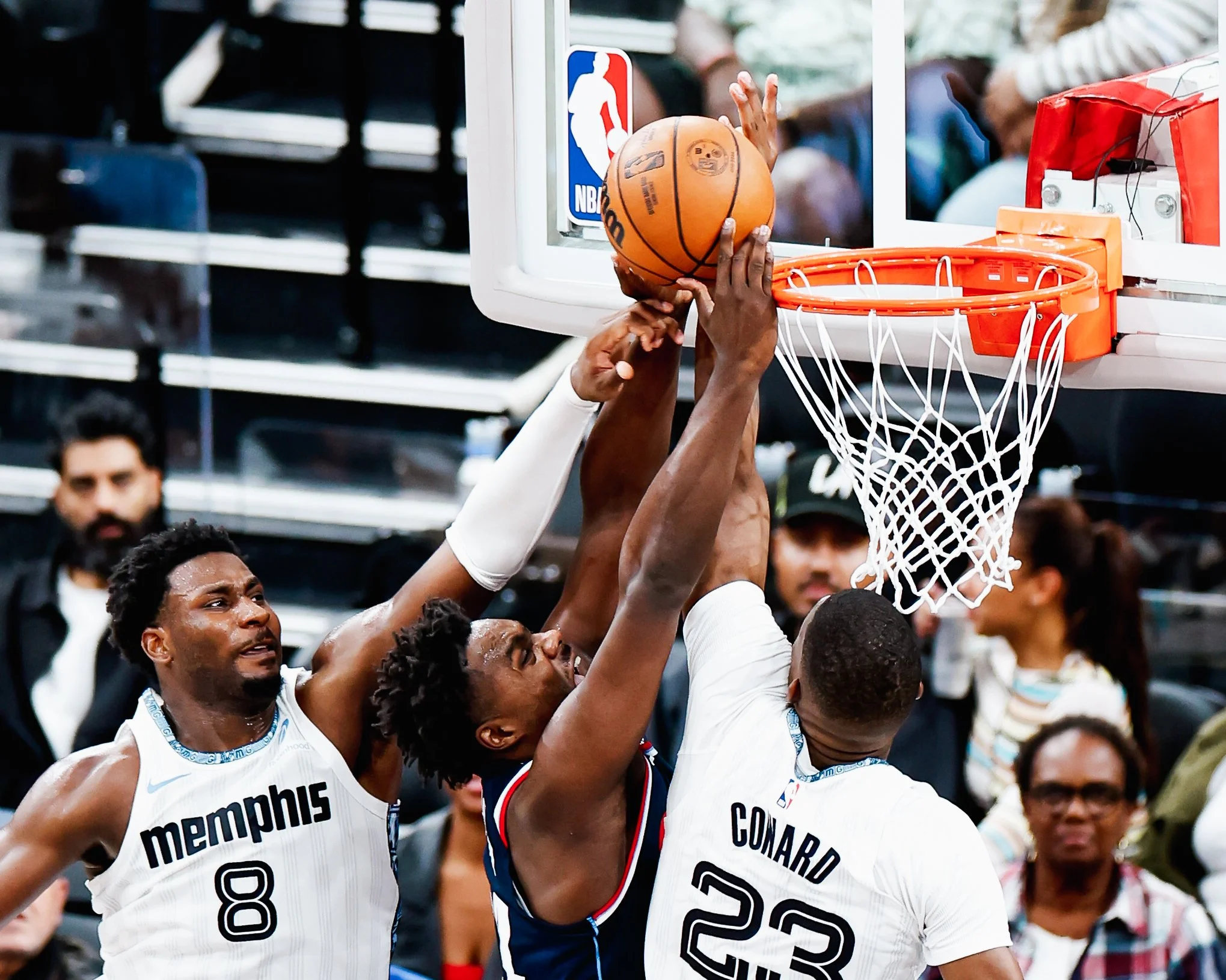 Three basketball players jump for a rebound near the basket, with one player attempting a shot while two others defend. The scene is in an indoor arena with spectators in the background.