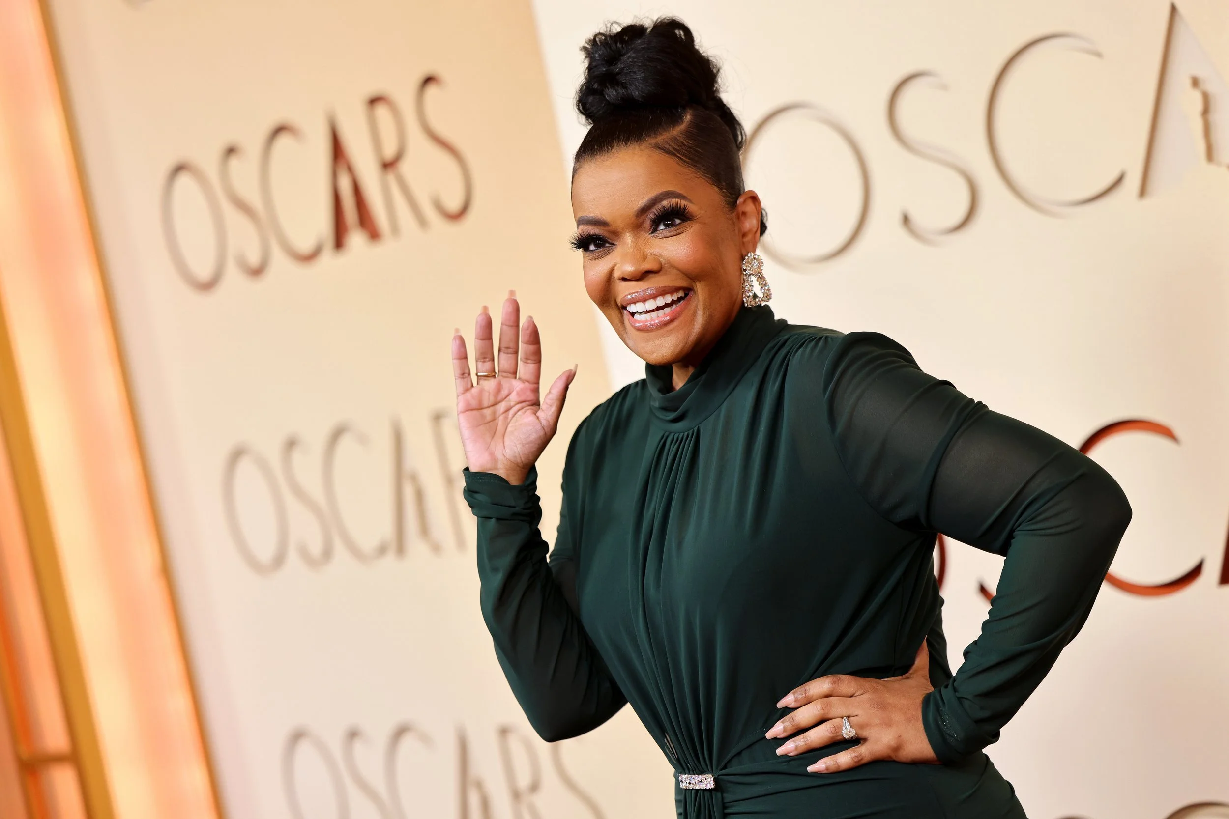 A woman in a dark green dress smiling and waving at the Oscars event.