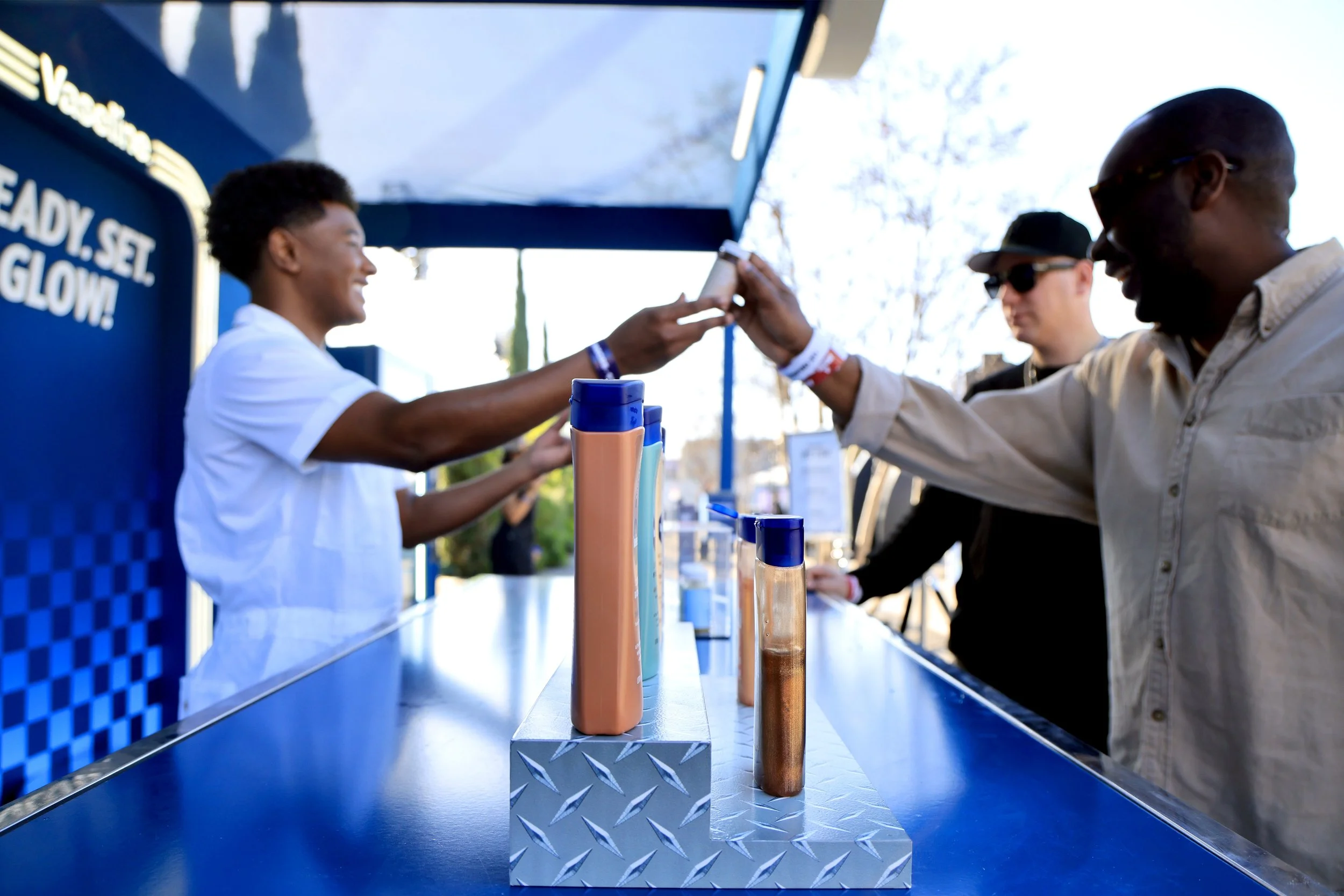 A man at a beverage stand handing his card to a smiling woman worker as he places an order during the daytime. Other people wait in line behind him, with a blue sign with white text that says "Ready. Set. Glow!" in the background.