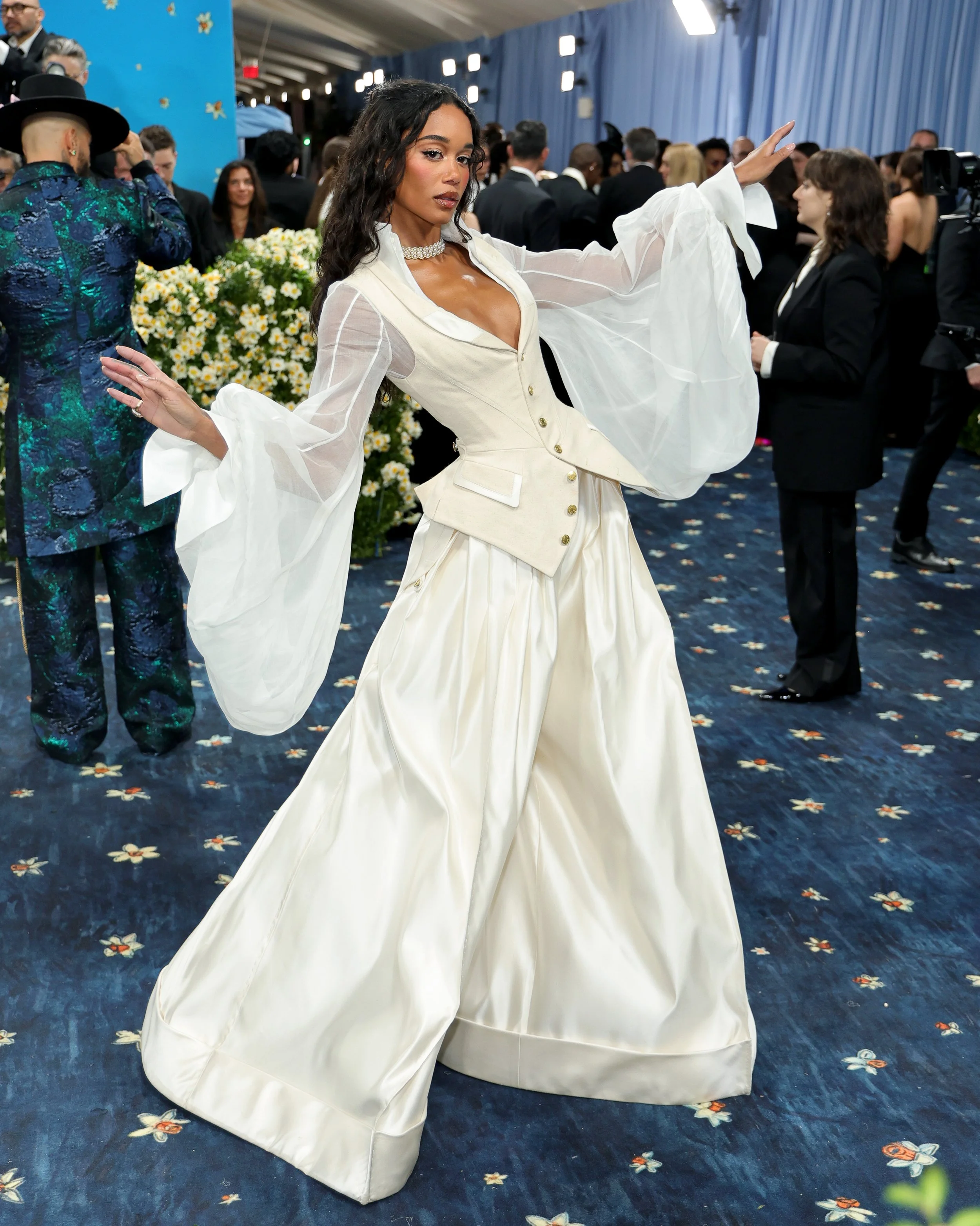 A woman in a vintage cream-colored dress and sheer white sleeves posing at a formal event with other attendees in the background.