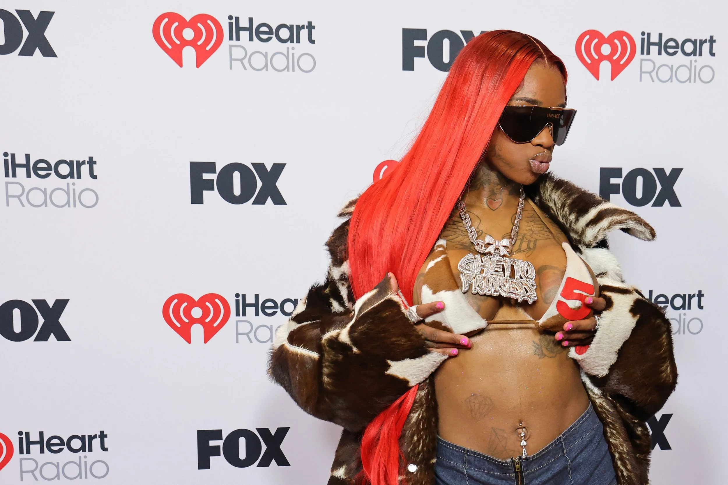 A woman with bright red hair, wearing sunglasses and a fur coat, poses on the red carpet at a Fox and iHeartRadio event.