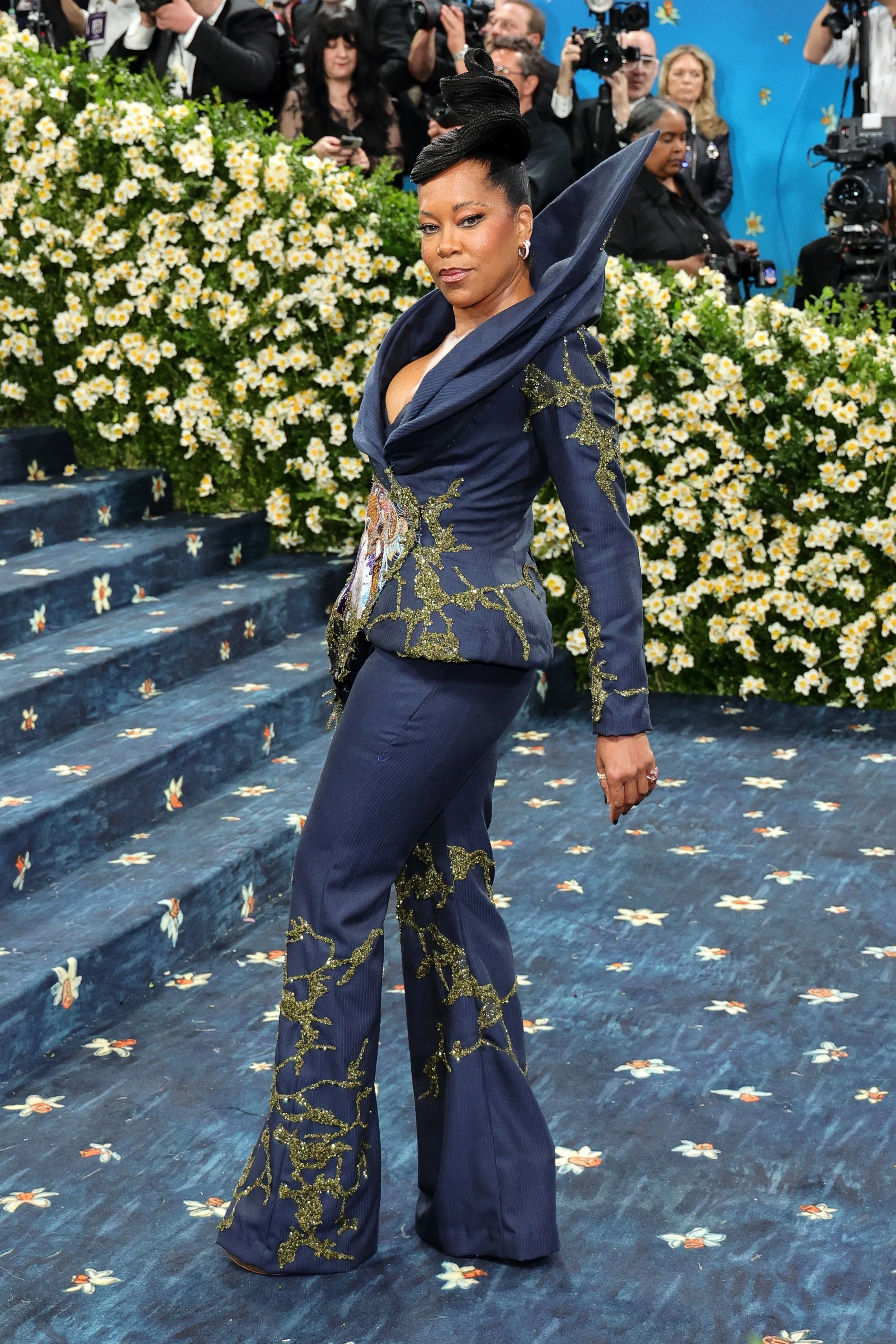 A woman in a dark blue, intricately embroidered suit with gold accents, standing on a floral-patterned carpet at a formal event, surrounded by photographers and other attendees.