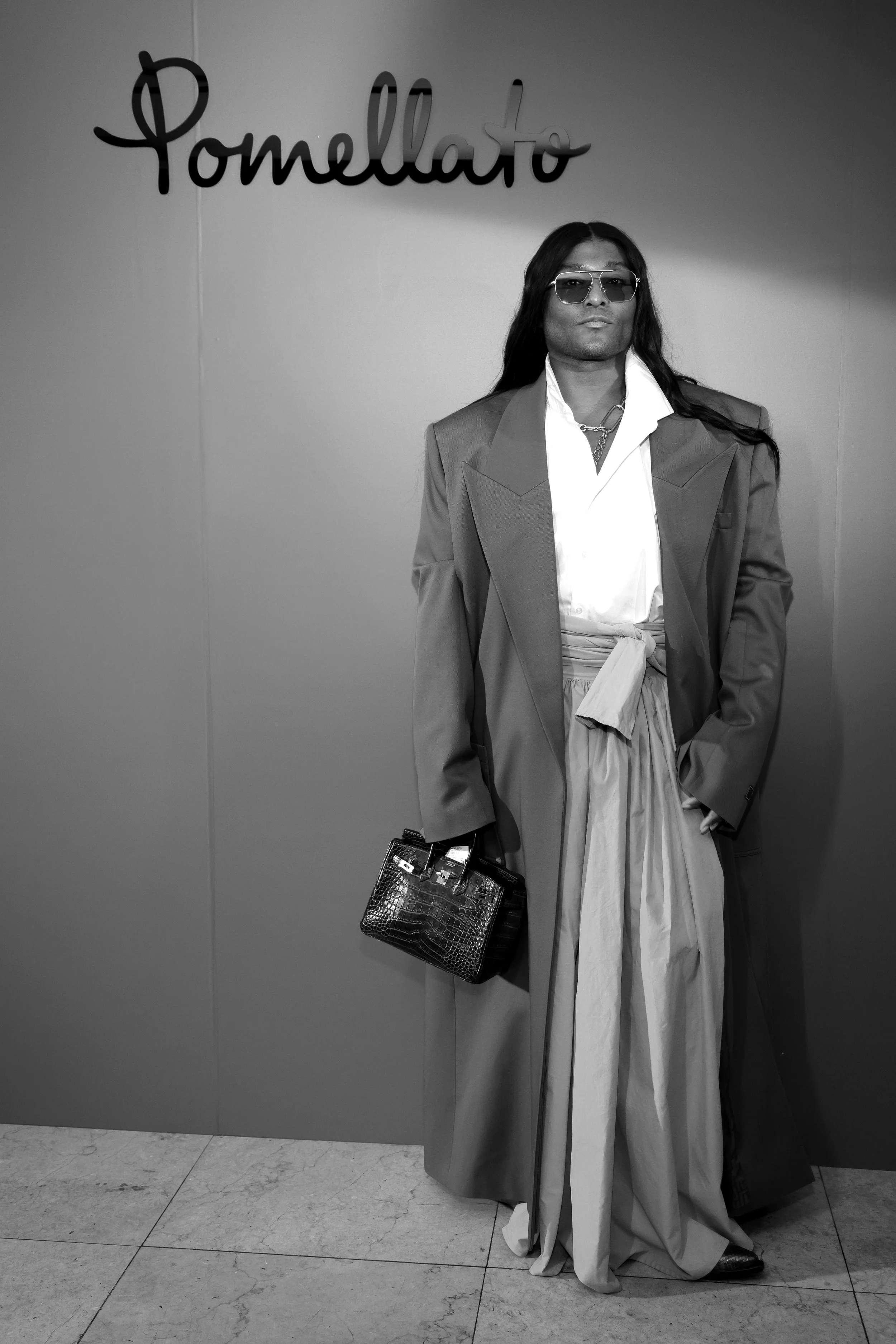 A person standing against a wall with the word 'Pomegranate' written on it. The person is wearing sunglasses, a blazer, wide-legged pants with a tied waist, and is holding a textured handbag.