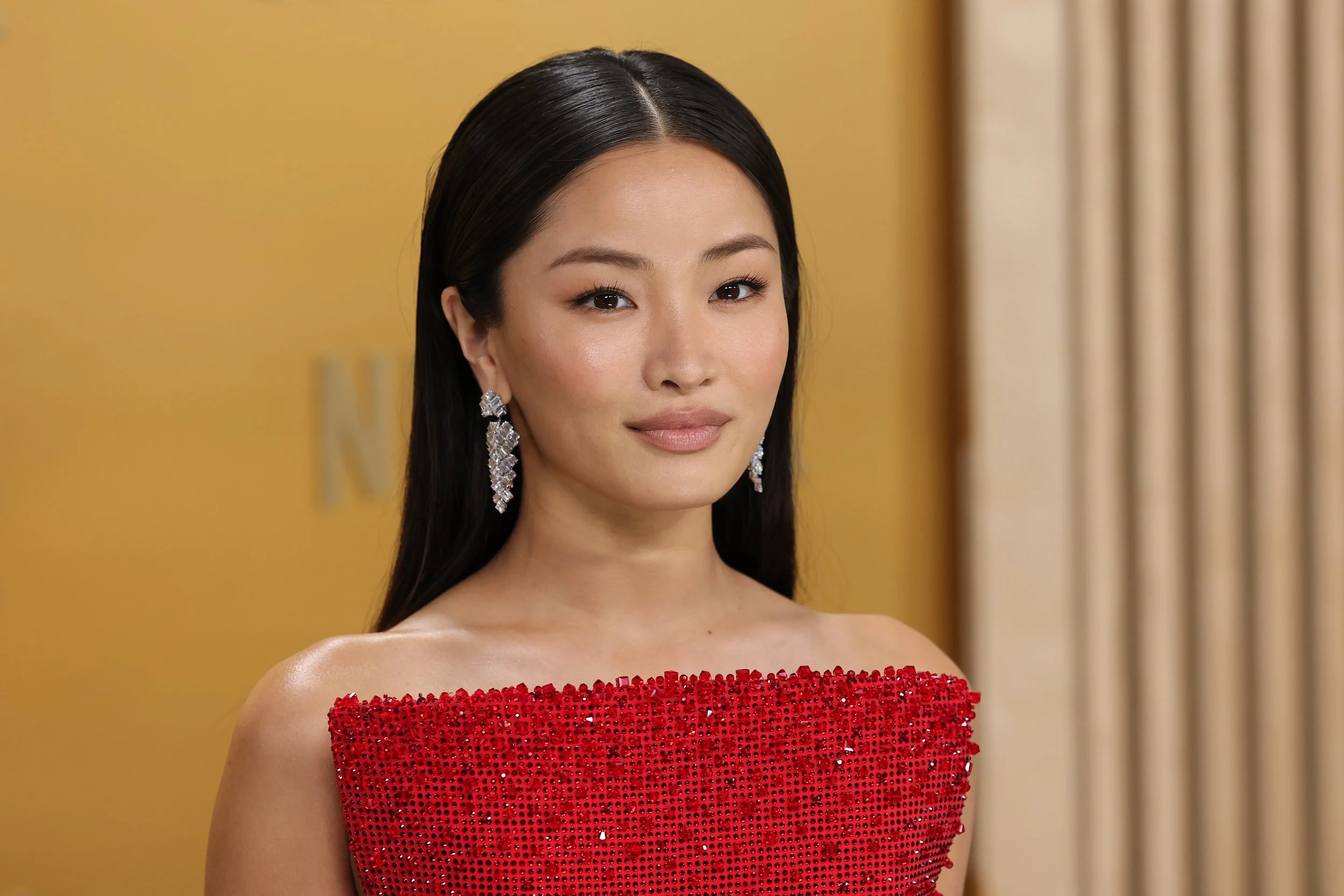 A woman with long black hair, wearing diamond earrings and a red off-shoulder, sequined dress, standing indoors with a blurred yellow background and beige curtains.