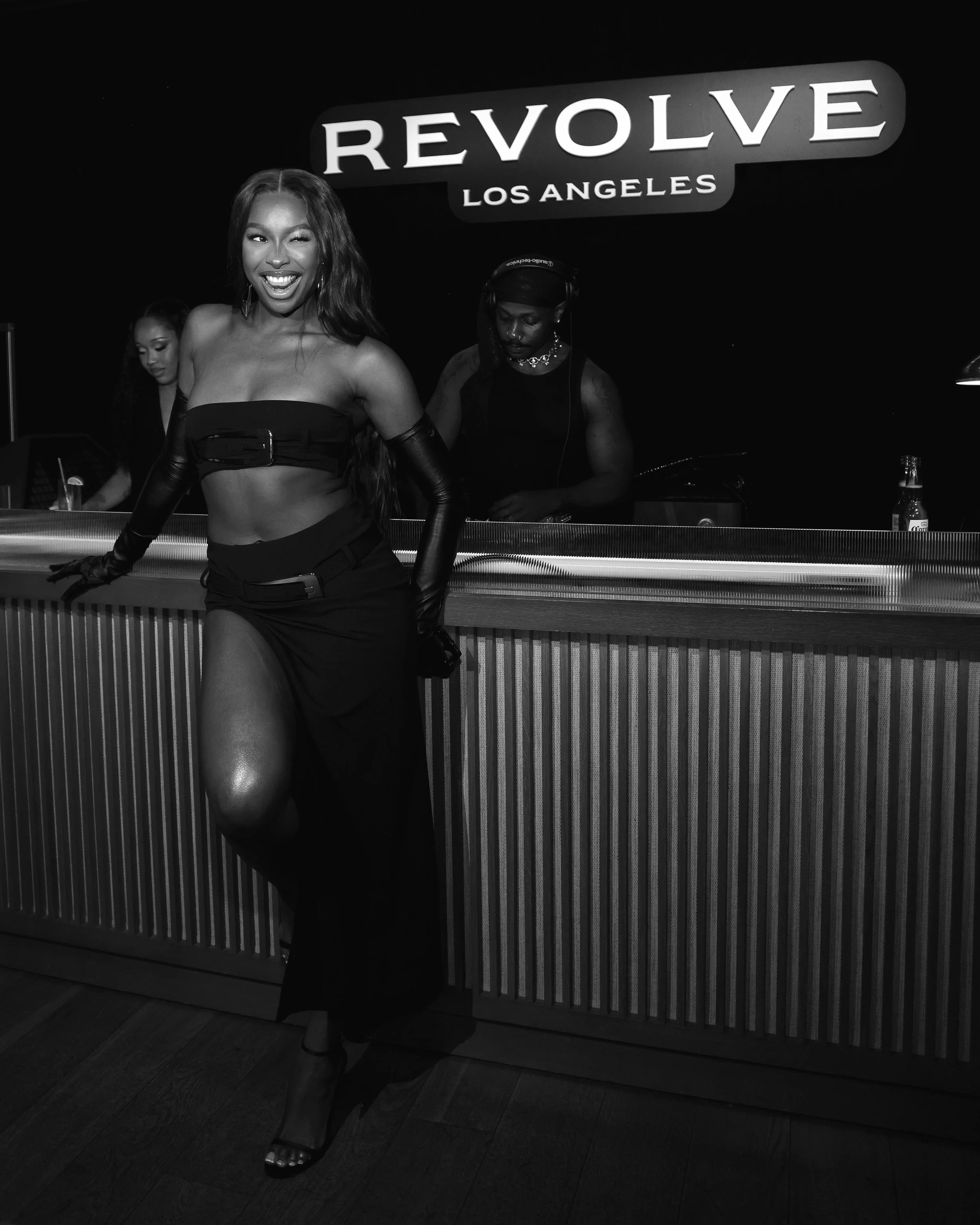 A woman in a strapless black crop top and long black skirt with a high slit, posing and smiling at a nightclub named Revolve Los Angeles, with two women behind a counter.
