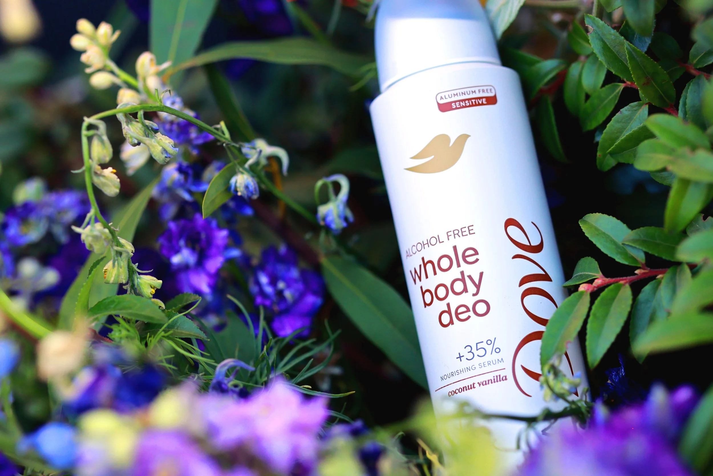 A can of alcohol-free whole body deodorant with coconut vanilla scent, surrounded by green leaves and purple flowers.