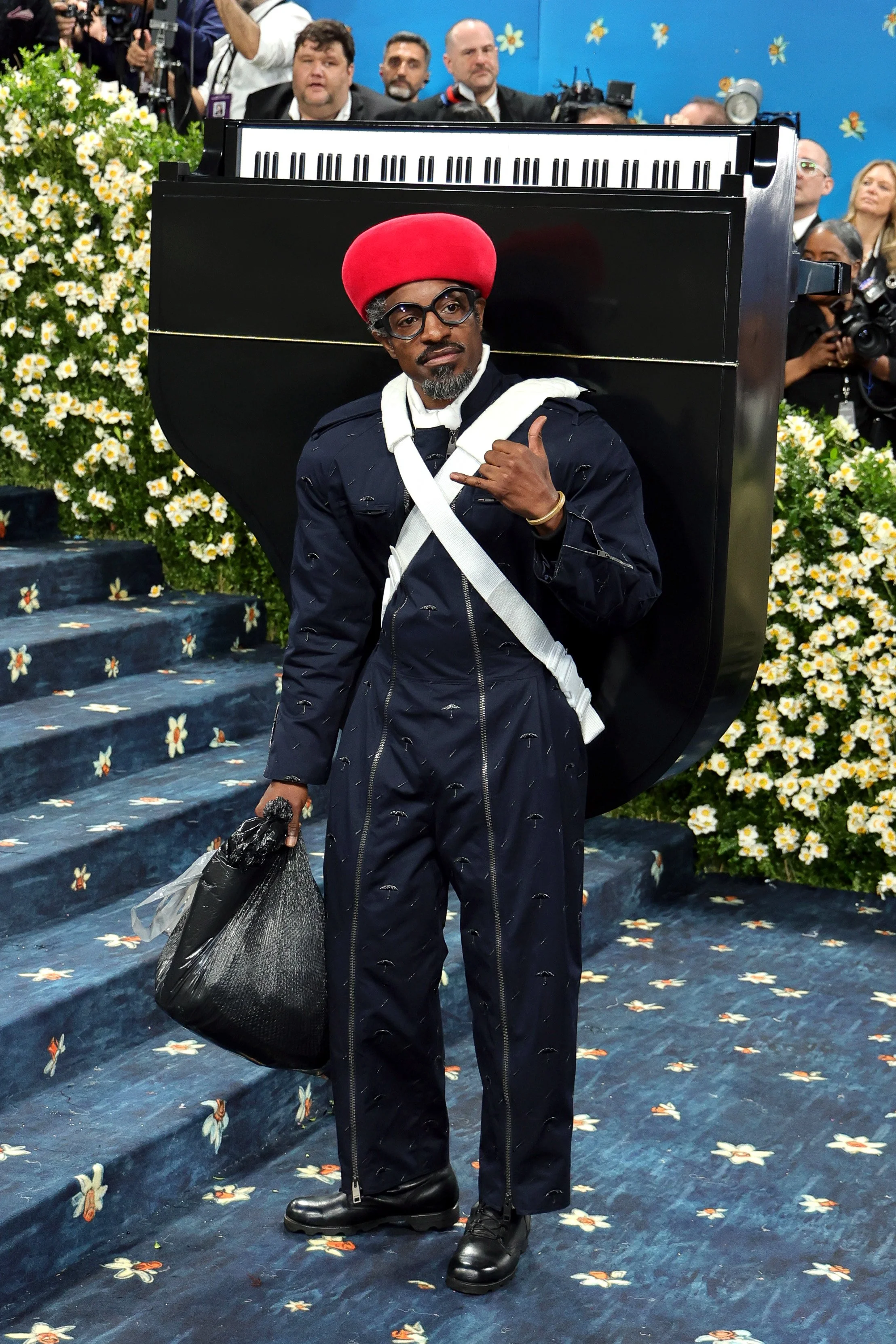 A man dressed in a dark suit with a pattern of small umbrellas, wearing glasses and a red hat, stands on stairs decorated with flowers. He is carrying a black bag in his left hand and making a hand gesture with his right hand. Behind him, there is a 