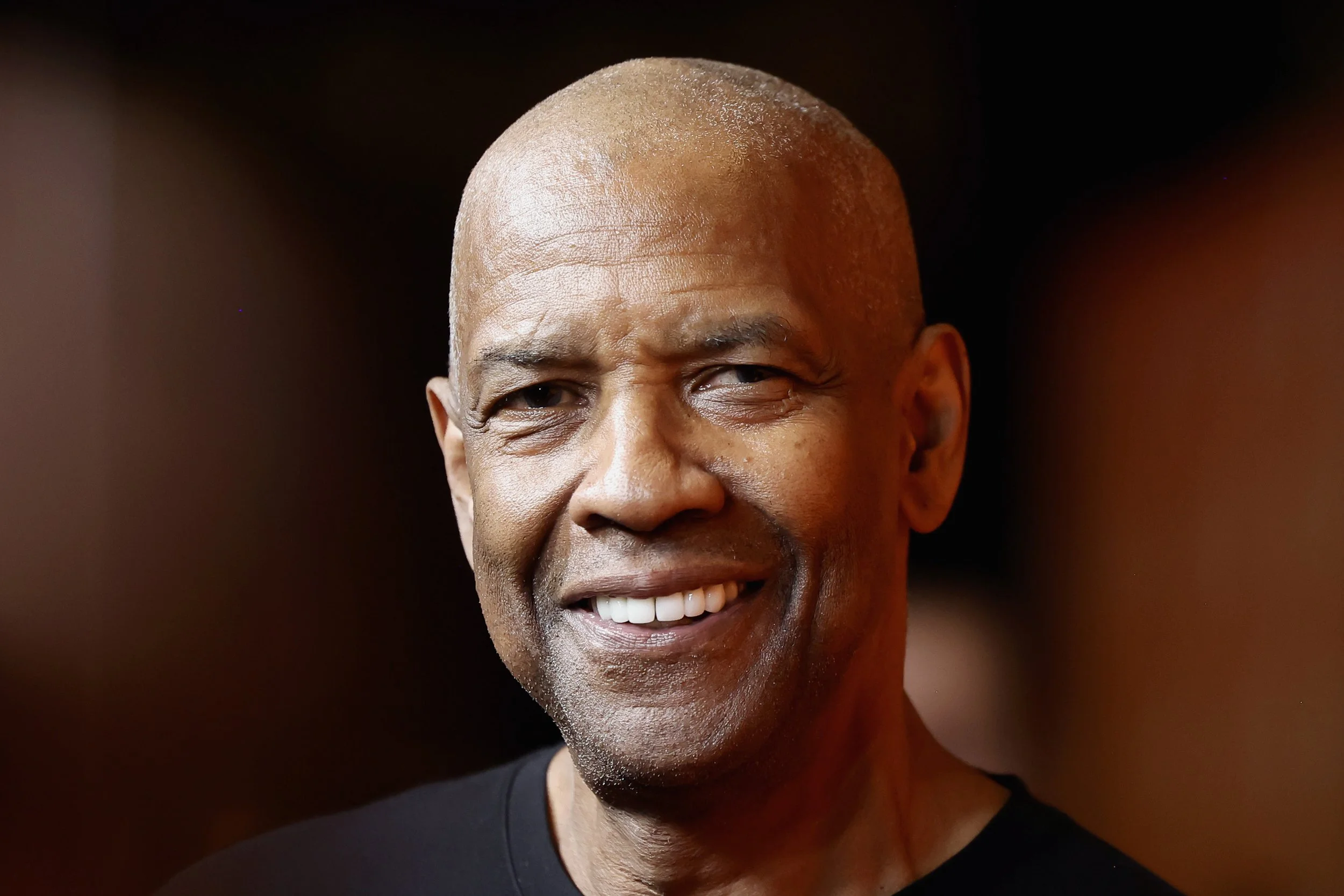 Close-up of a smiling middle-aged man with a bald head and dark shirt, with a blurred dark background.