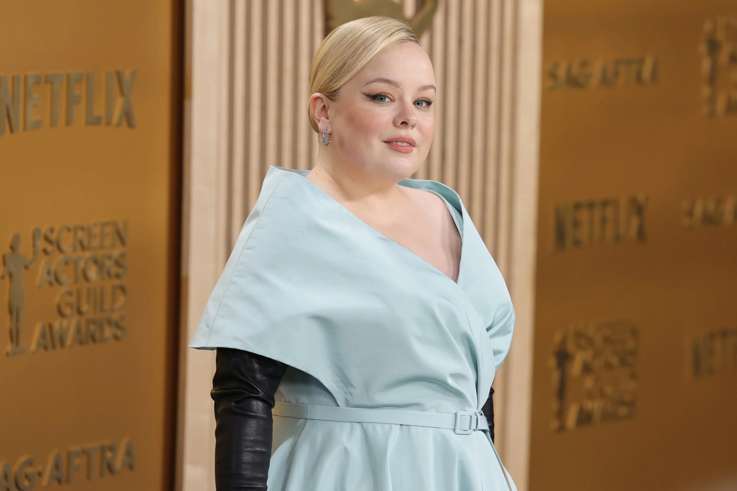 A woman with blonde hair wearing a light blue dress with black sleeves standing in front of a beige curtain at an award event.