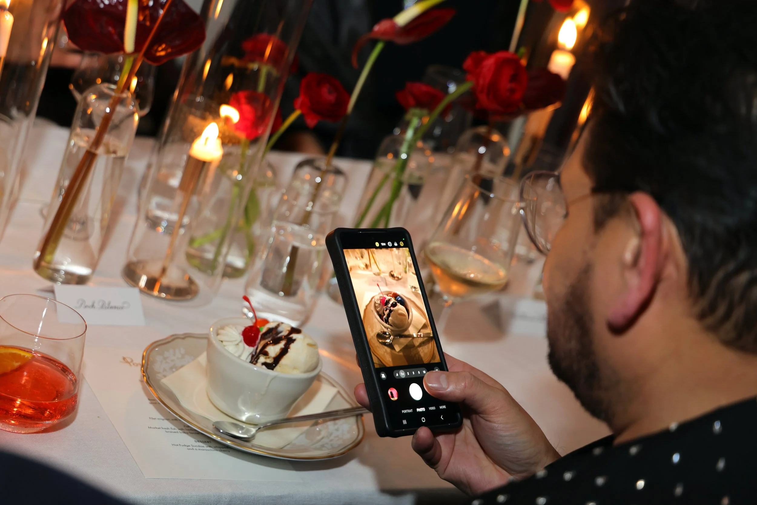 A person taking a photo of a dessert with a smartphone at a decorated table with flowers and candles