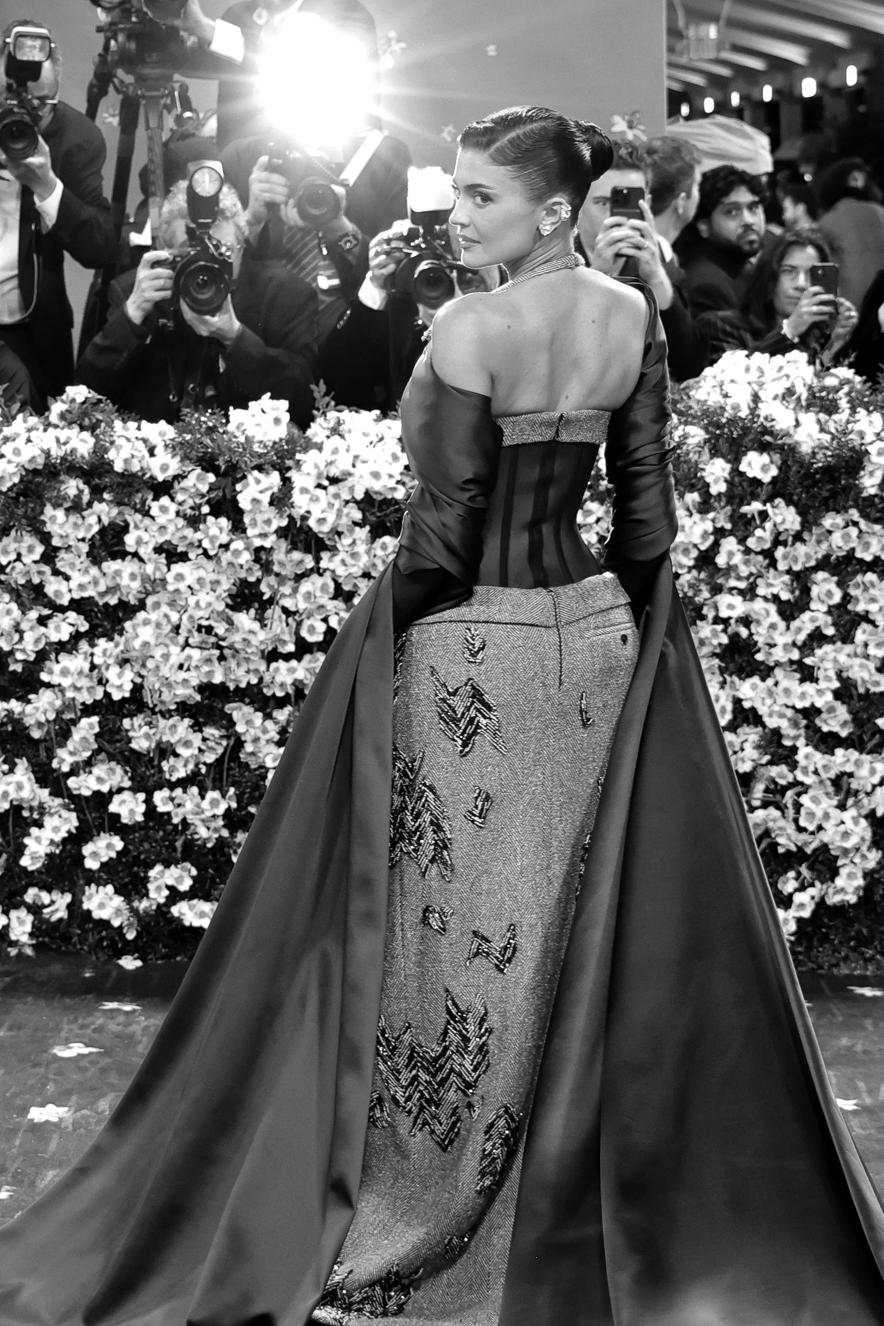 A woman in an elegant gown on a red carpet, with photographers taking pictures and a floral backdrop.