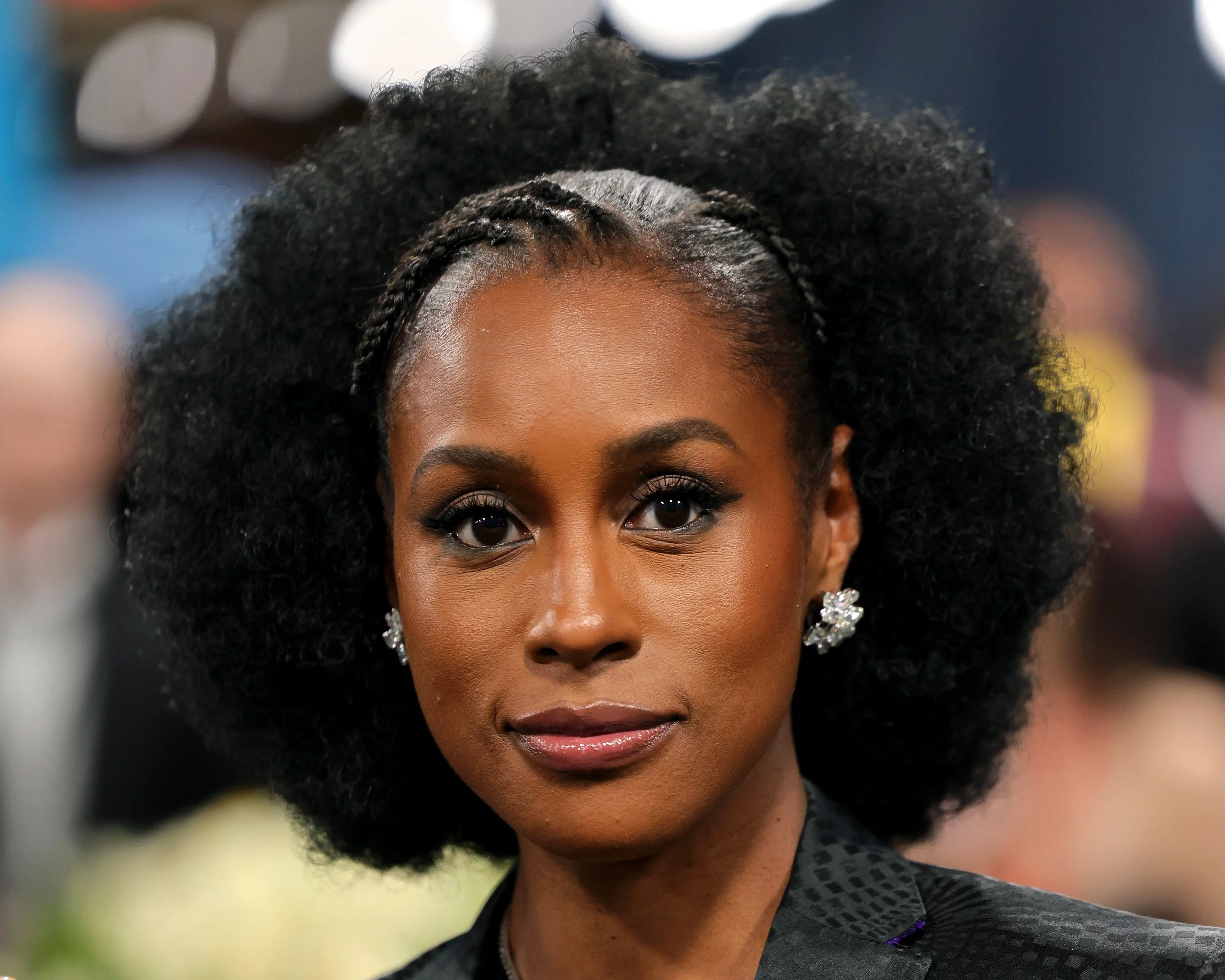 Close-up of a woman with dark skin, curly afro hair with braided accents, wearing floral earrings and a textured dark blazer in a crowded indoor setting.