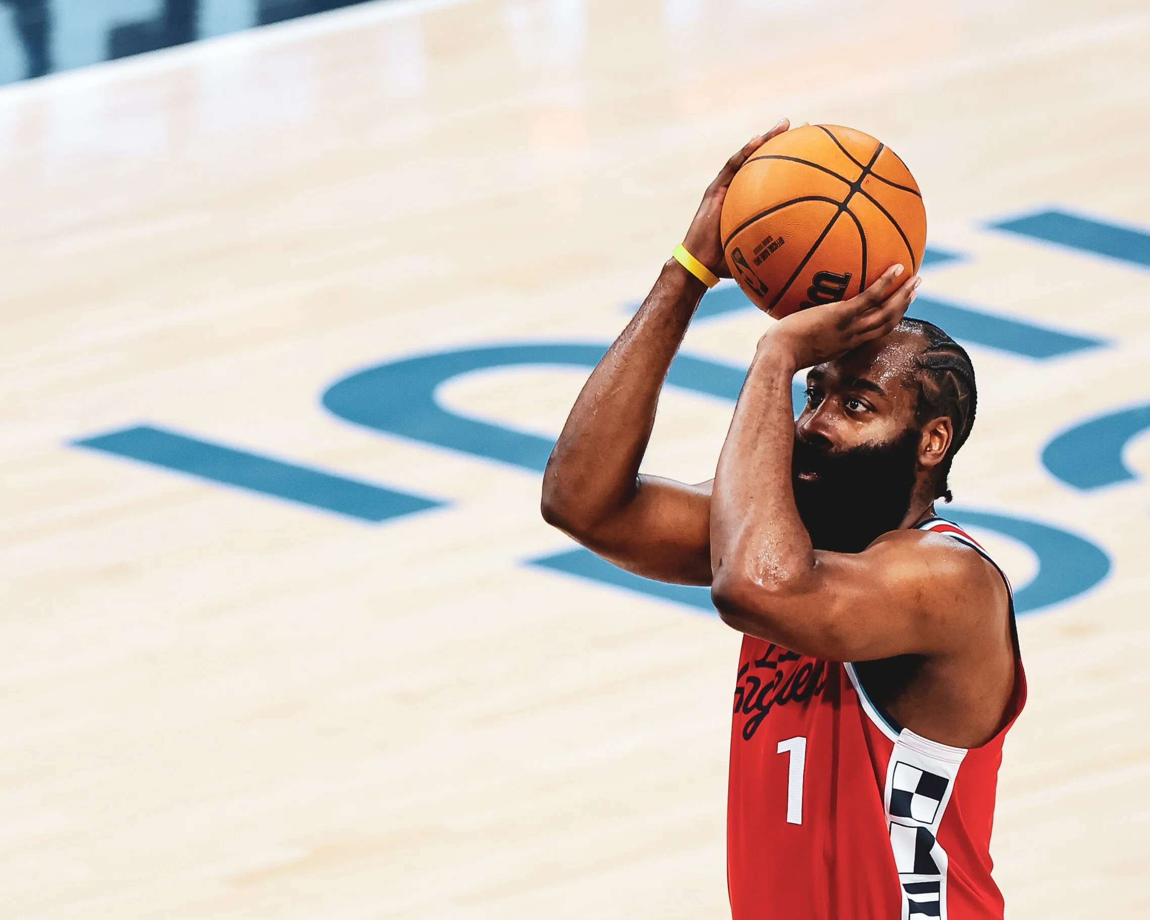A basketball player prepares to take a shot on the court, holding the basketball above his head with both hands, wearing a red jersey with the number 1.