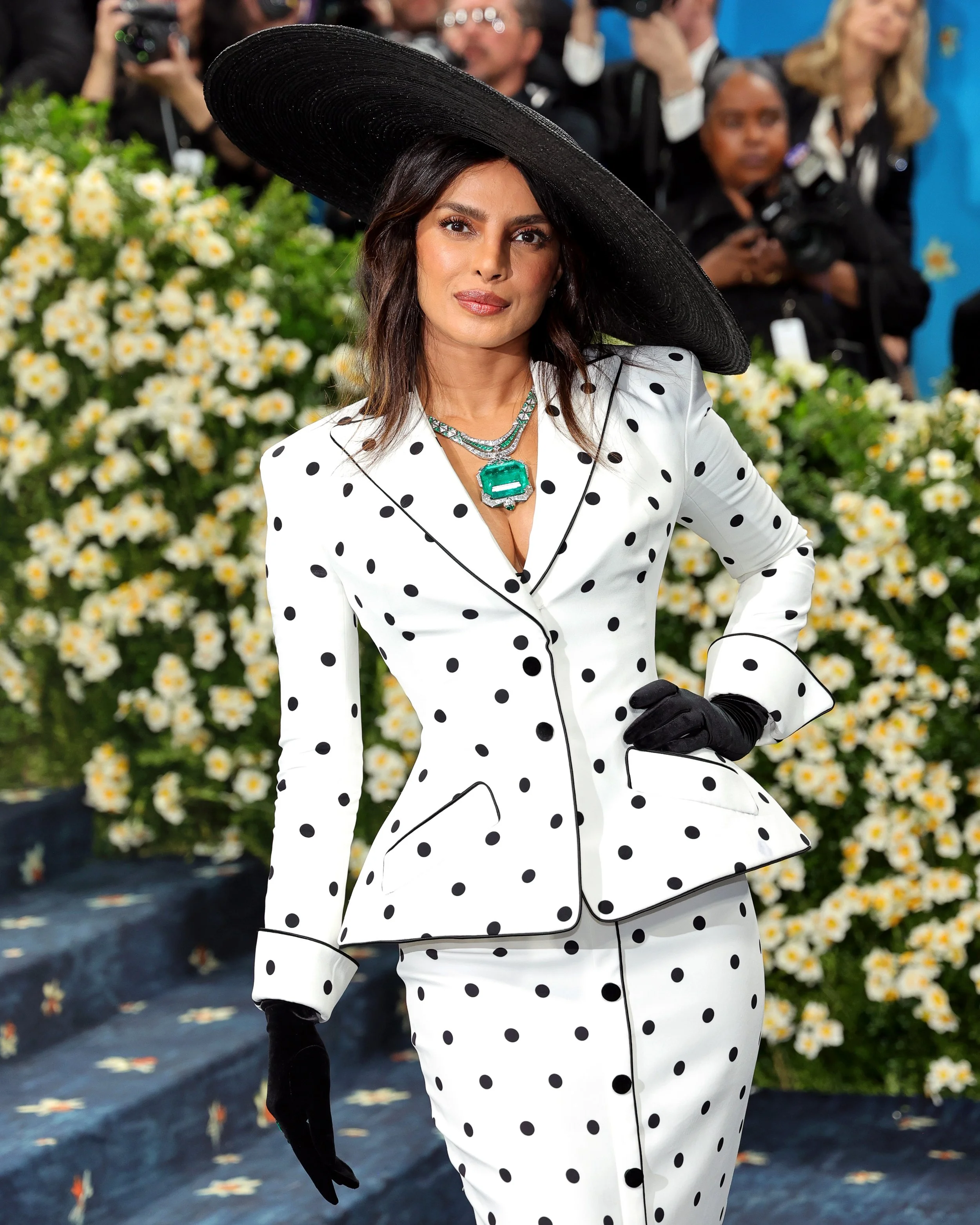 Woman wearing a black wide-brimmed hat, white suit with black polka dots, and black gloves, standing outdoors with a background of white flowers and people taking photos.