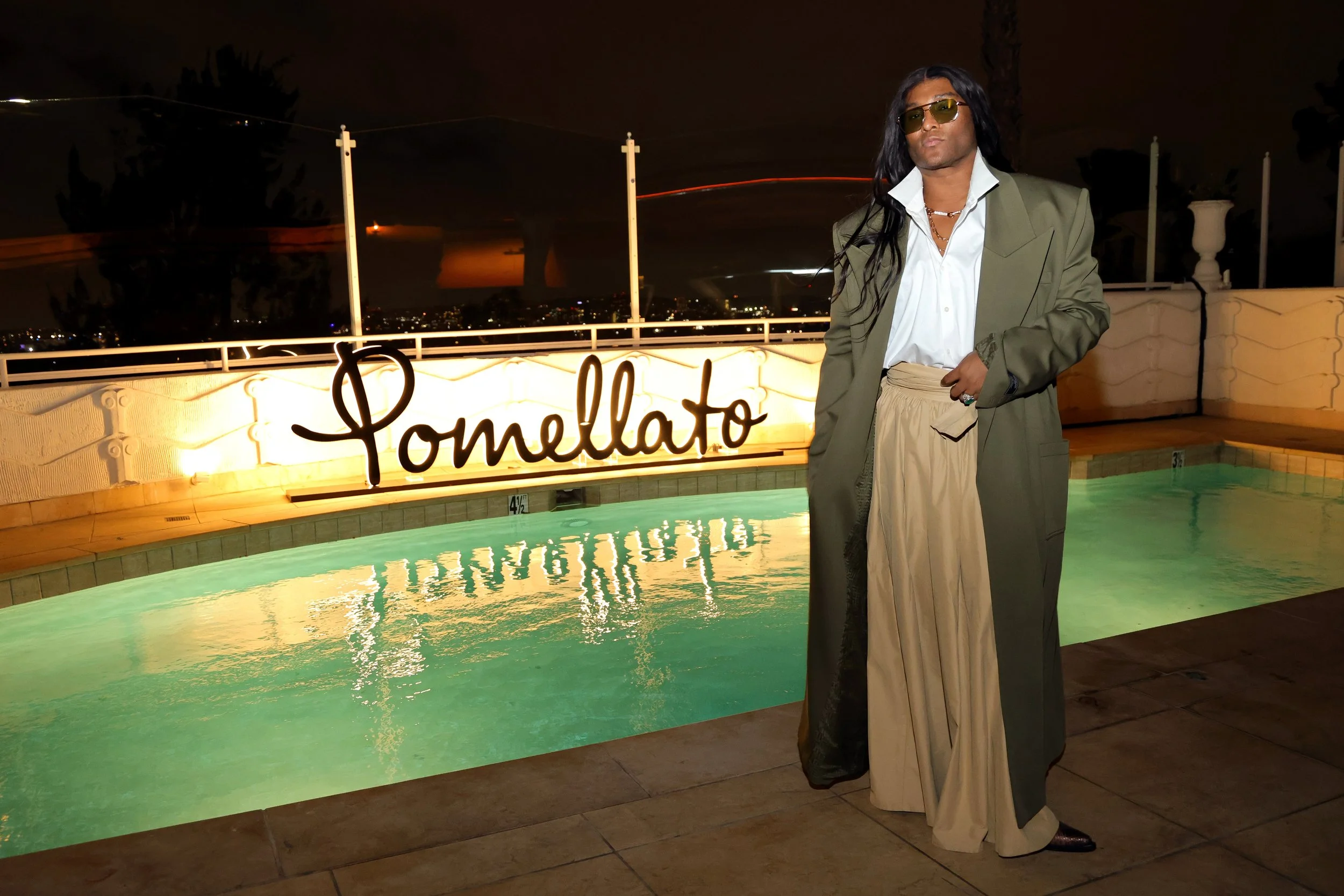 A person standing next to a lit pool with a sign that reads 'Pomellato' at night, wearing sunglasses and an oversized suit with long hair.
