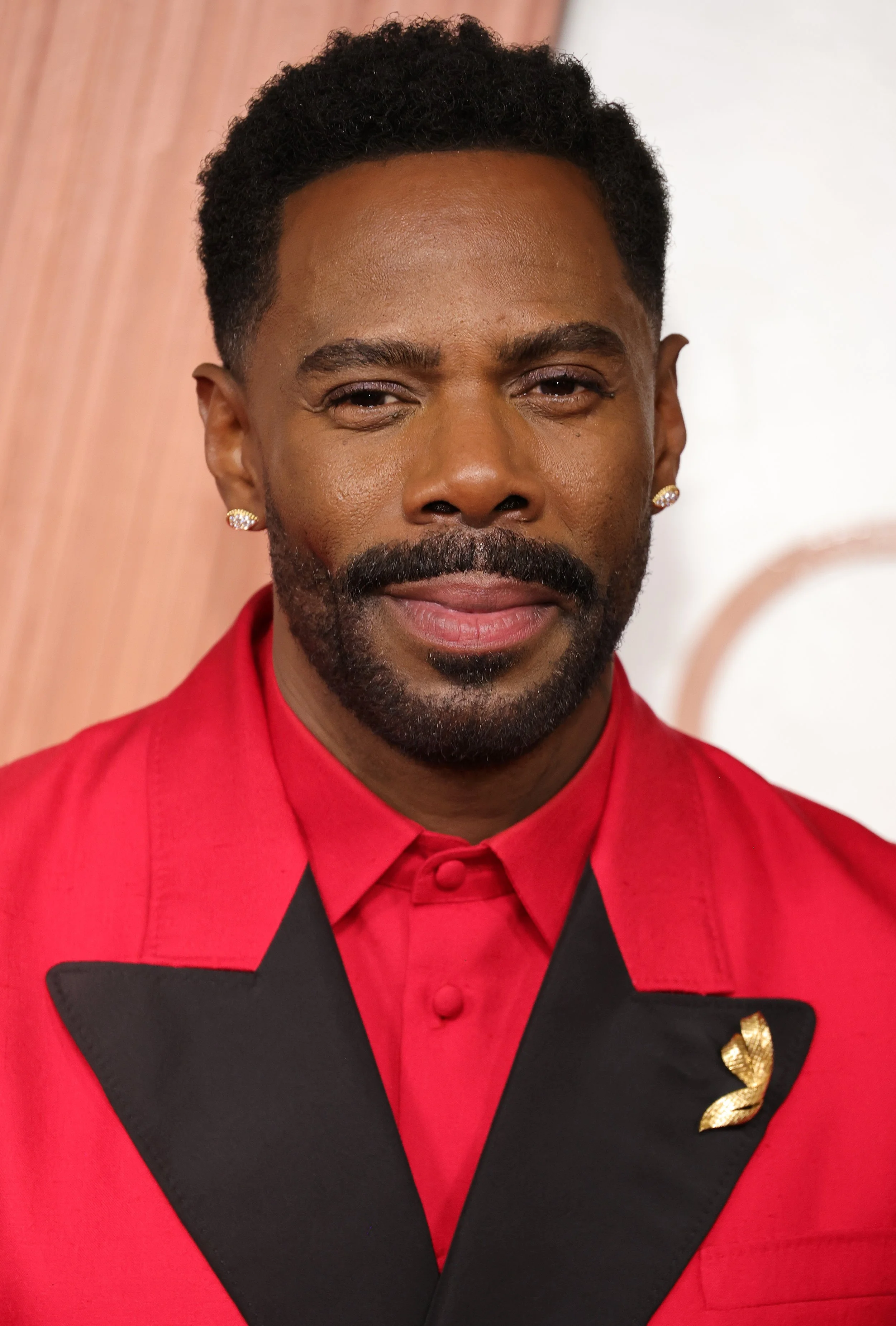 A man with dark skin, wearing a red jacket with black lapel, a red shirt, and gold earrings, standing in front of a background with warm tones.