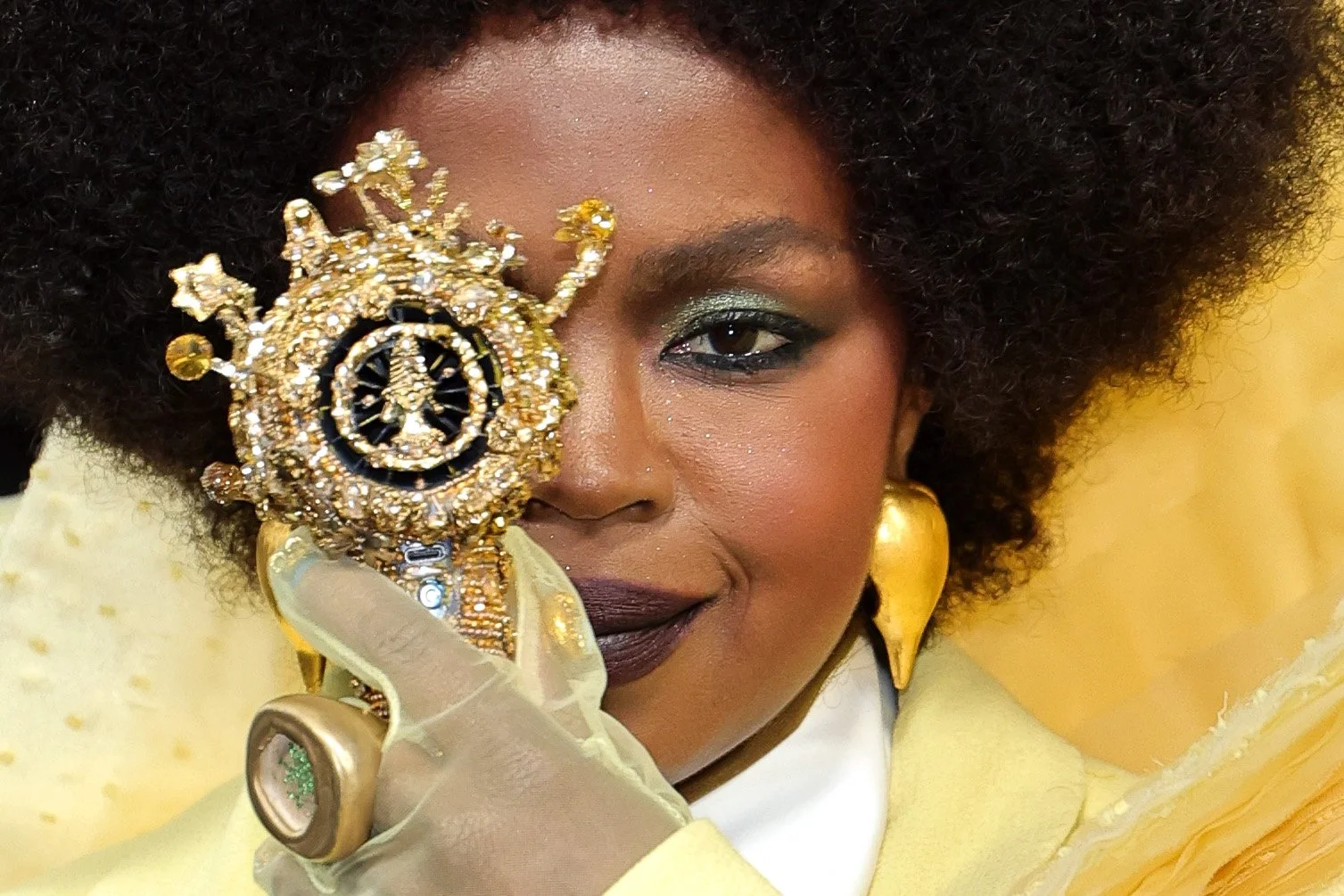 A woman with dark skin and curly hair holding an ornate gold and black circular object with jewelry, wearing gold earrings and a yellow garment, looking at the camera.